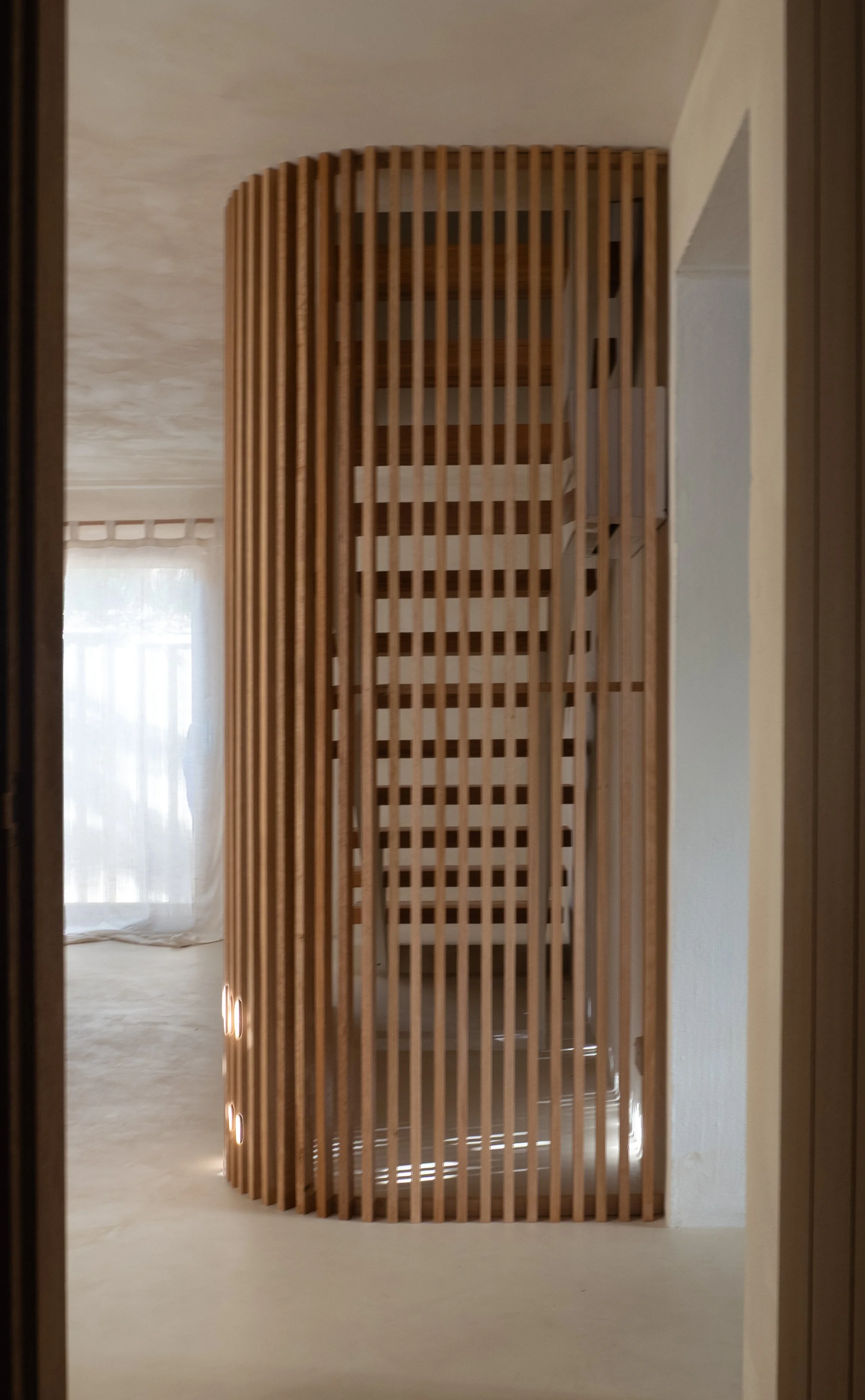Interior of a room with a curved wooden slat partition and a staircase visible in the background near a window with white curtains.