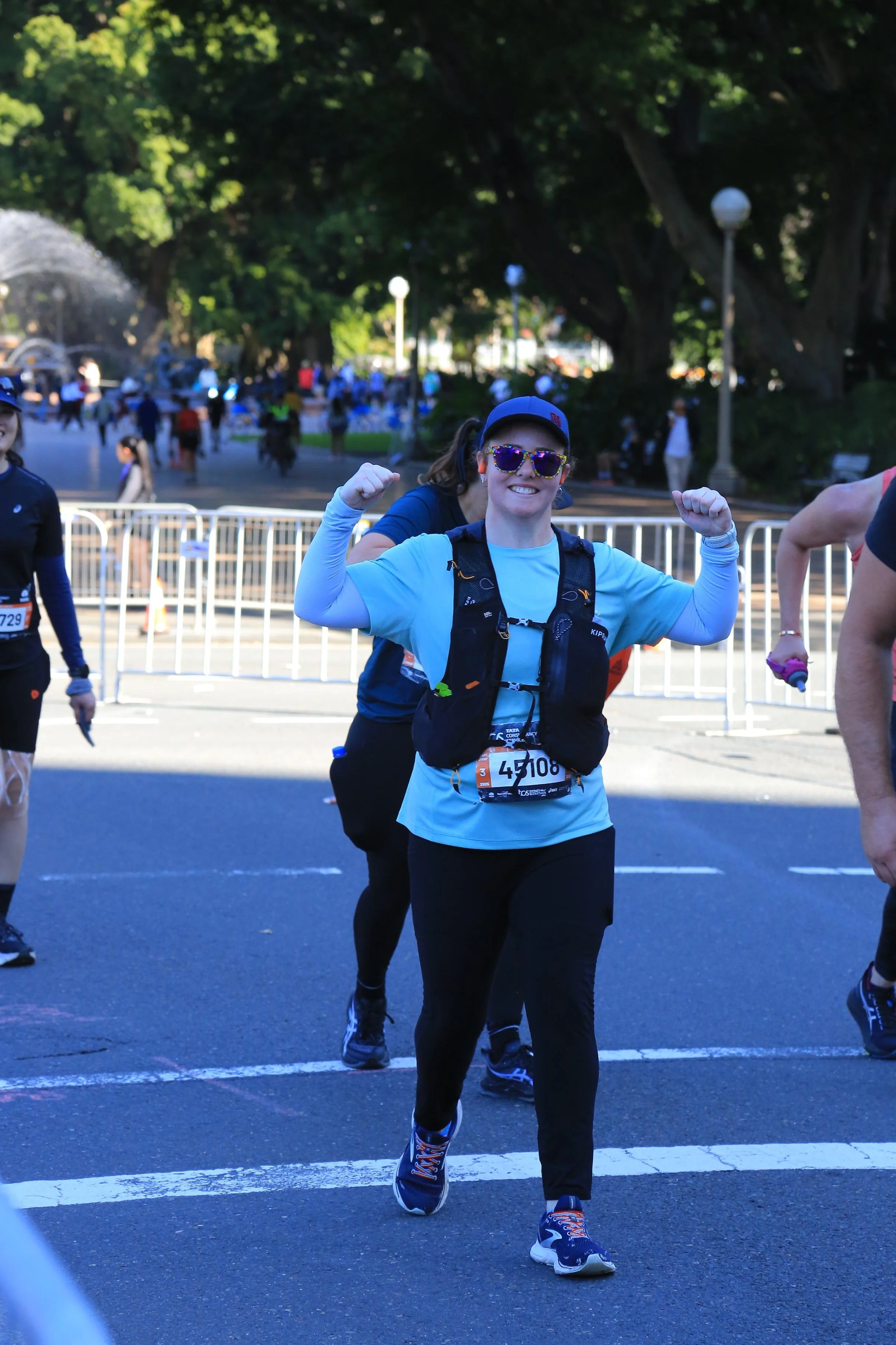 A woman wearing sunglasses and a blue shirt is smiling and flexing her arms. She is participating in a marathon or running event, with other runners visible in the background and race bib number 45108.