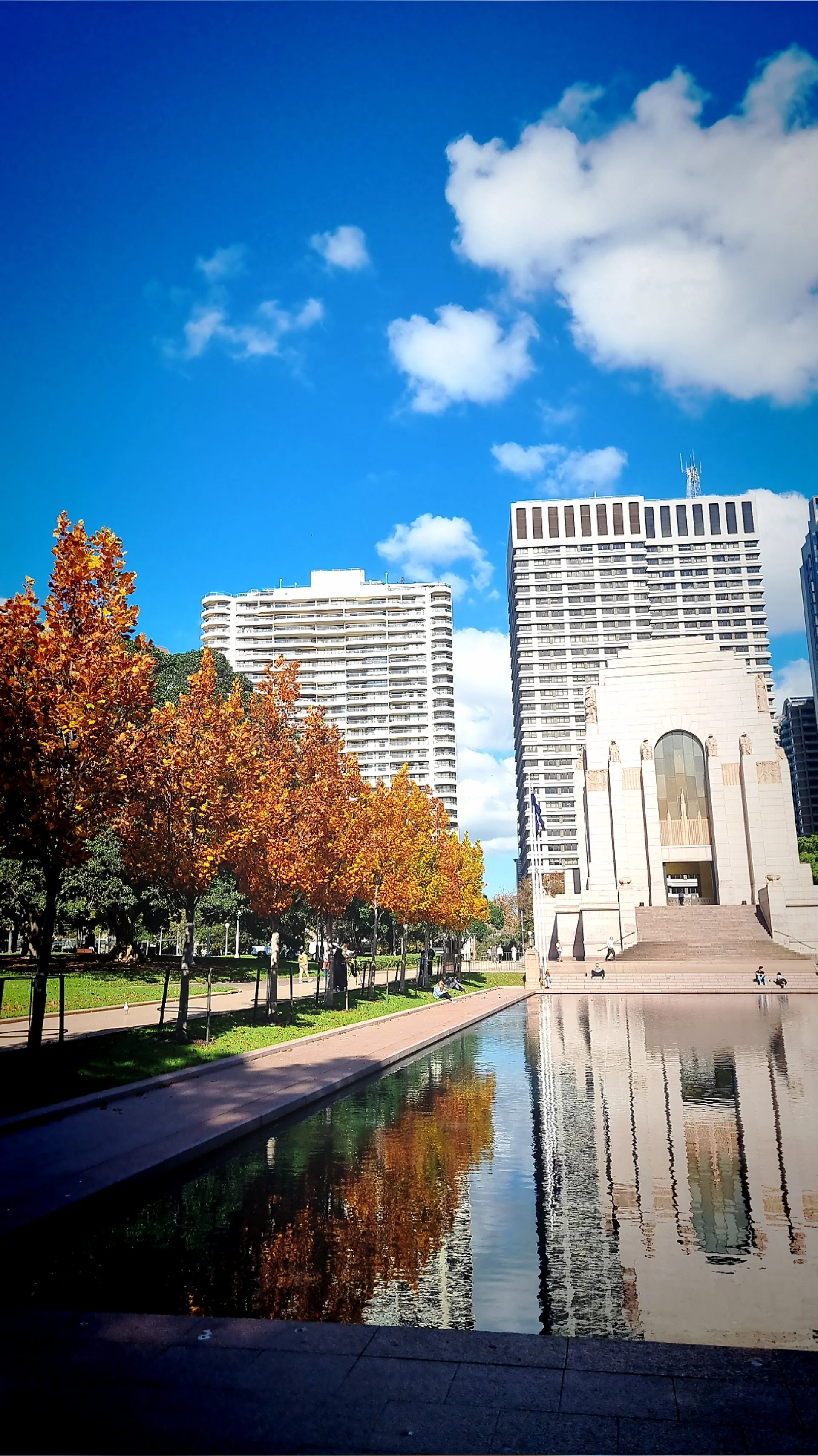 A city park with trees showing autumn foliage, a reflecting pool, and high-rise buildings in the background under a partly cloudy blue sky.