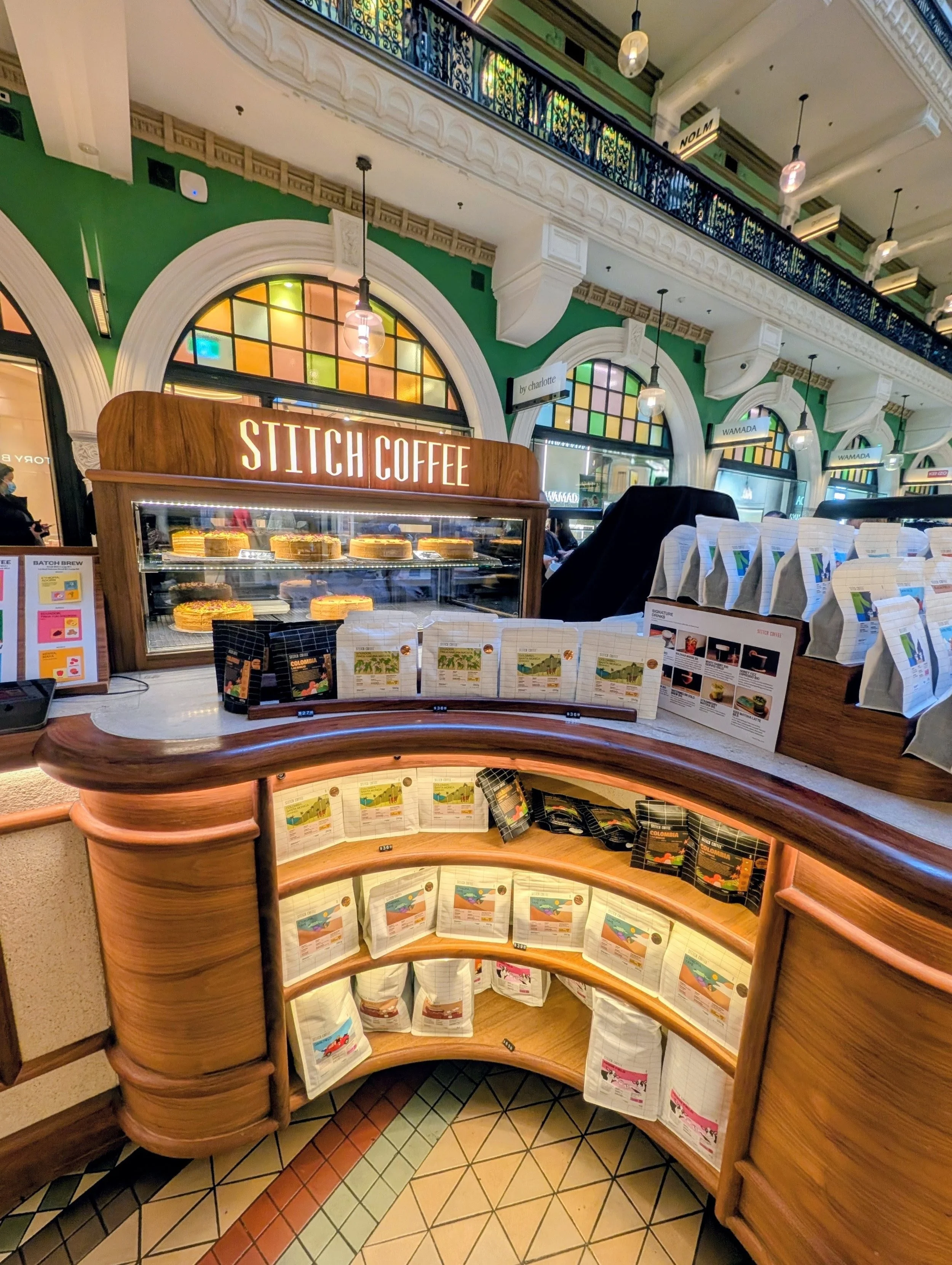 Indoor coffee shop with a wooden display case labeled 'Stitch Coffee' showcasing baked goods, green walls, colorful stained glass windows, and menu signs, with customers in the background.