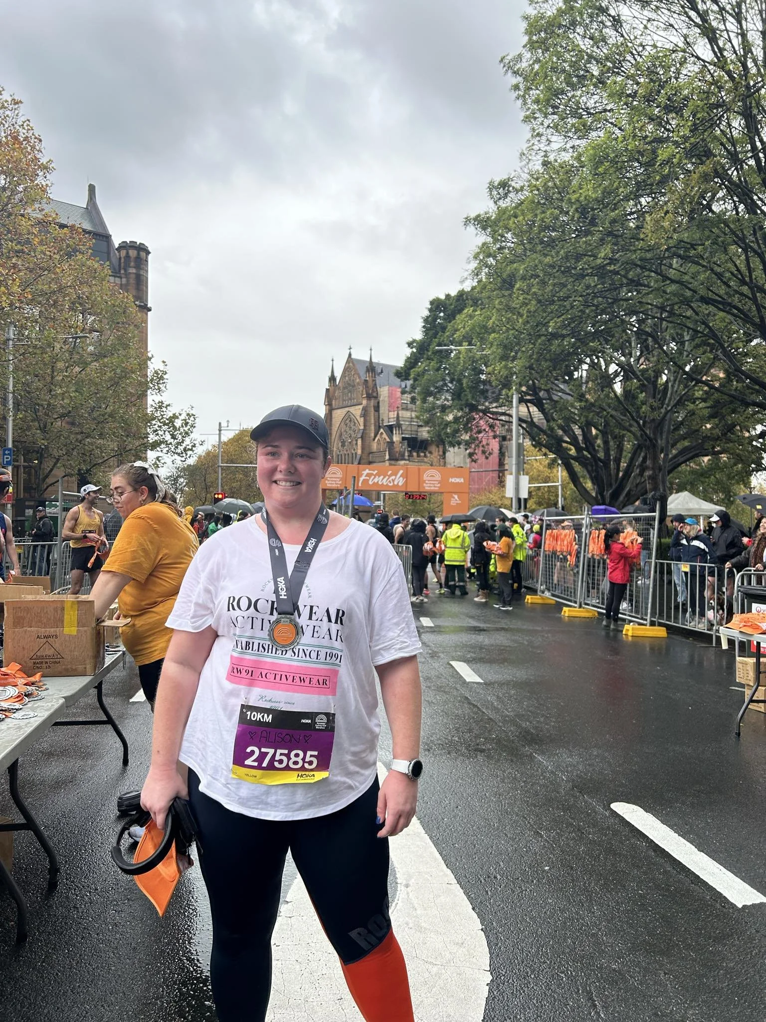 A woman with a race bib and medal smiling at the finish line of a marathon, with other runners and spectators in the background on a rainy day.