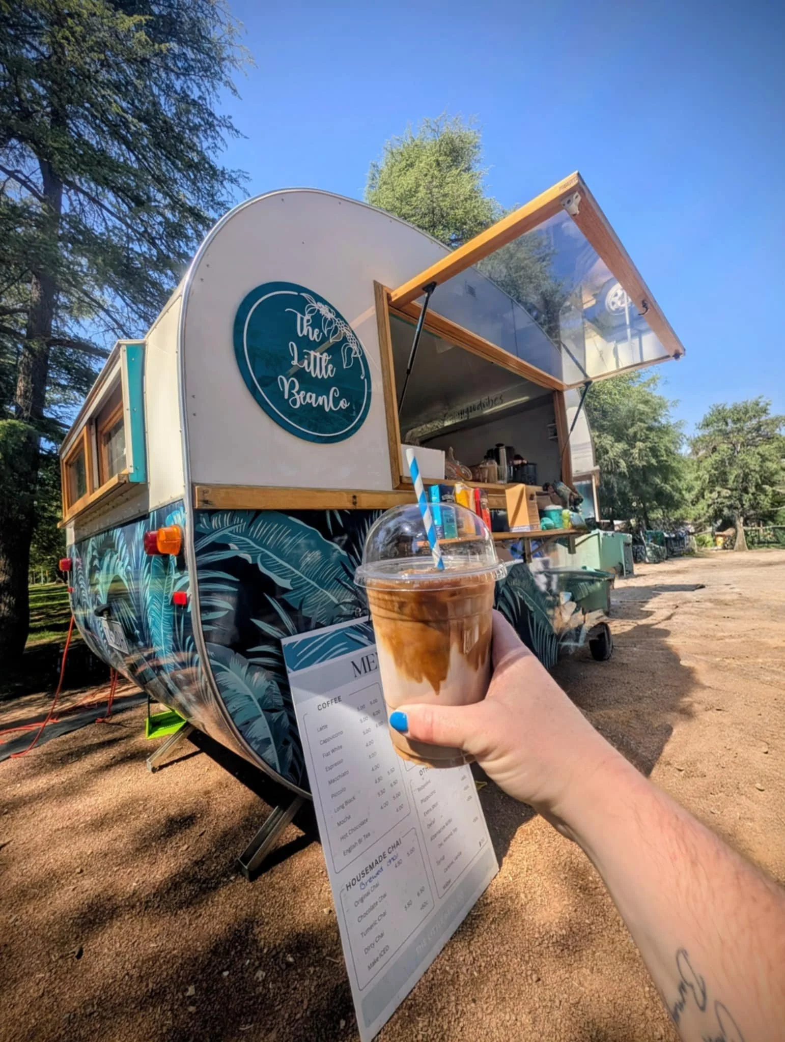 A person holding an iced coffee drink in front of a food truck called The Little Bean. The truck is decorated with tropical leaves and has a menu attached.