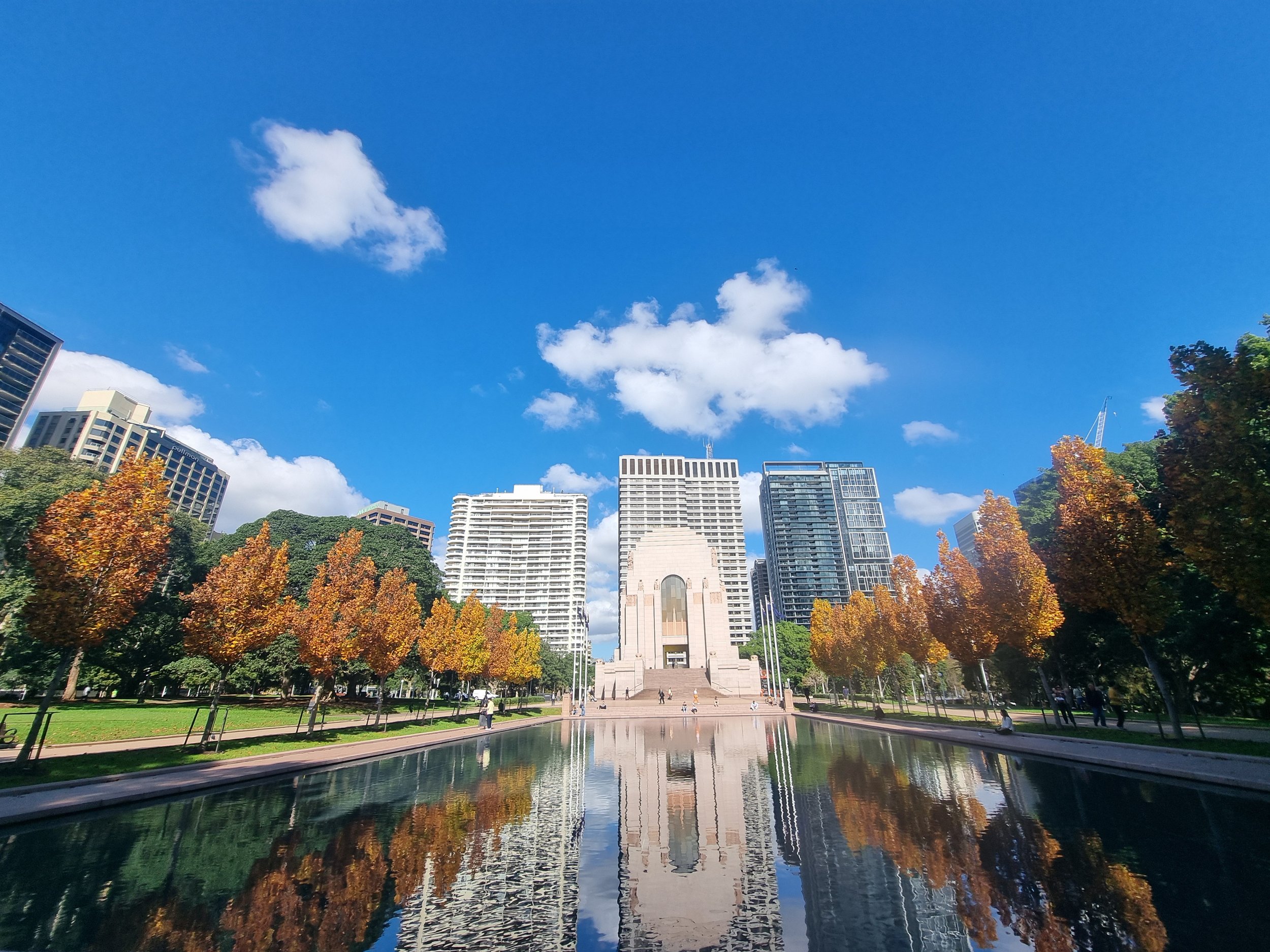 City park with trees in fall colors, a reflecting pool, and tall modern buildings in the background under a bright blue sky with a few clouds.