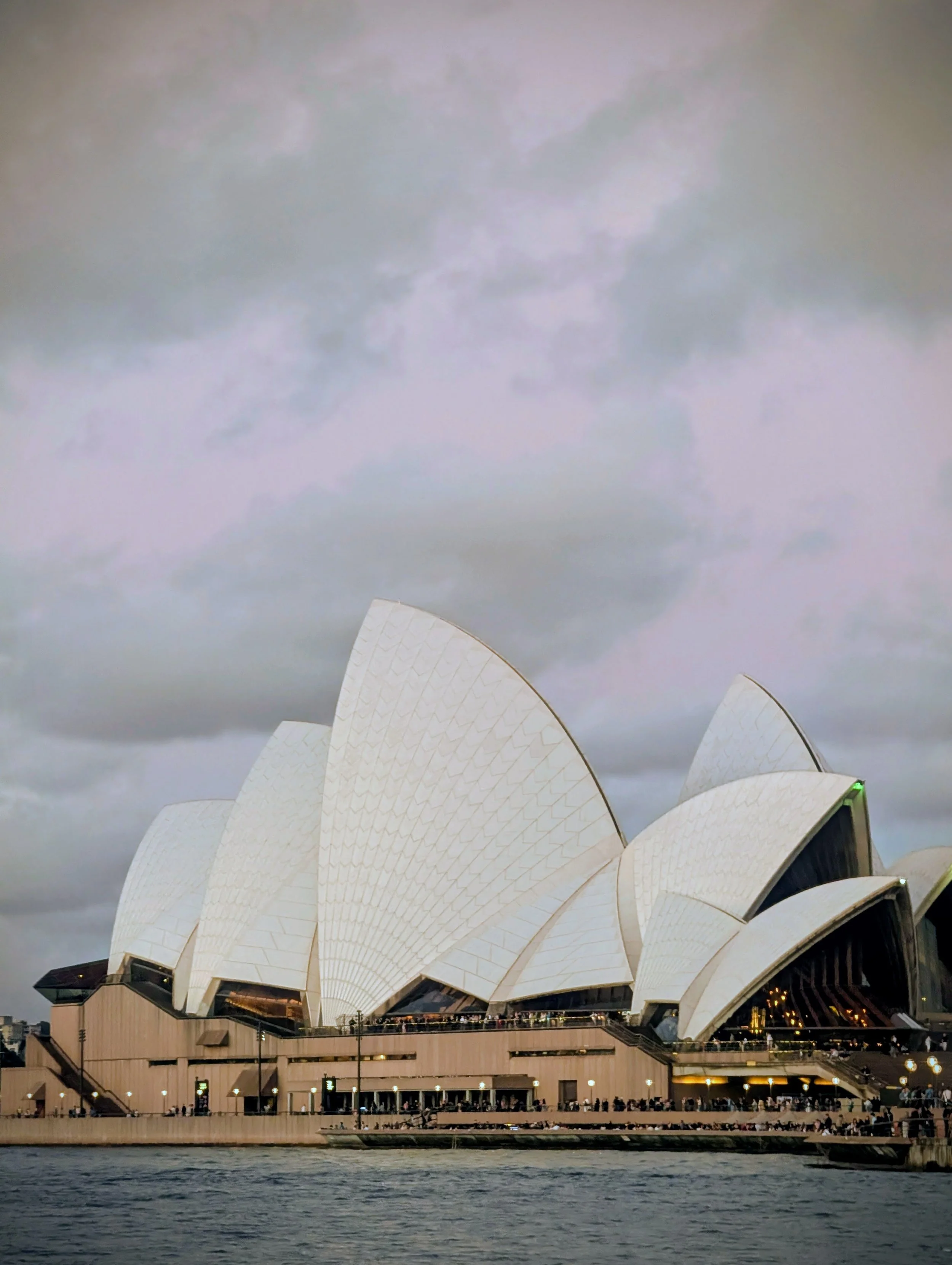 Night view of Sydney Opera House with lit-up sails, reflecting lights on the water in the foreground.
