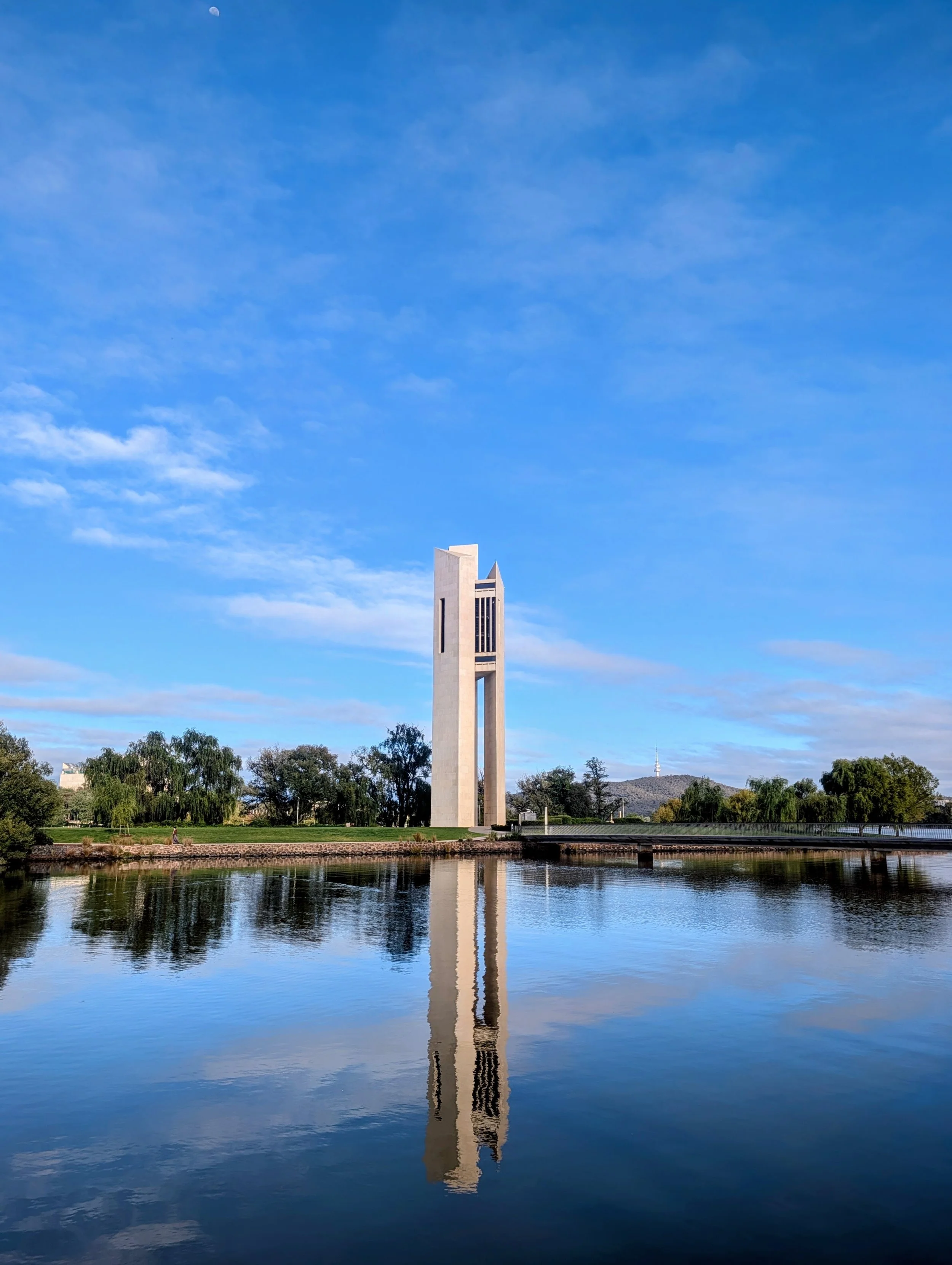 A tall, modern white monument reflecting in a calm body of water with trees and a hilly landscape in the background, under a blue sky with scattered clouds.