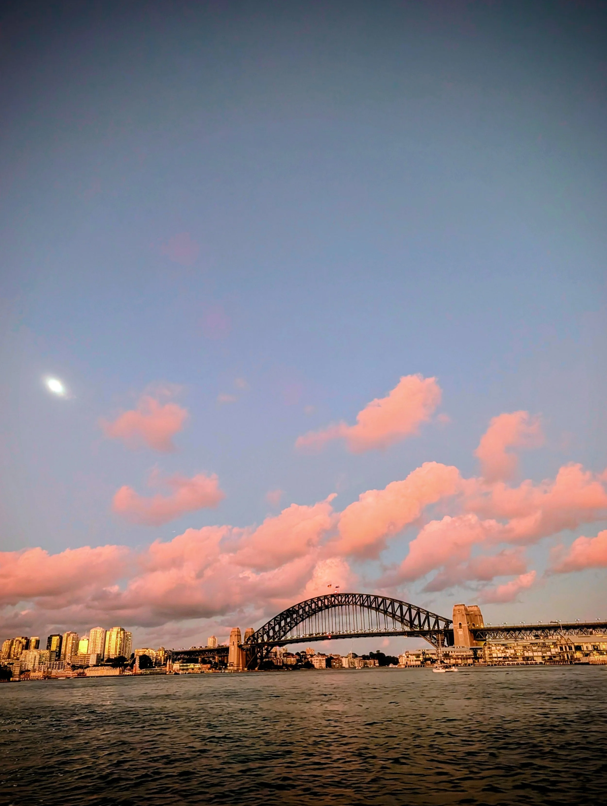 A city skyline with tall buildings, a large arch bridge, and a sky filled with pink and blue clouds at sunset, seen from across a body of water.