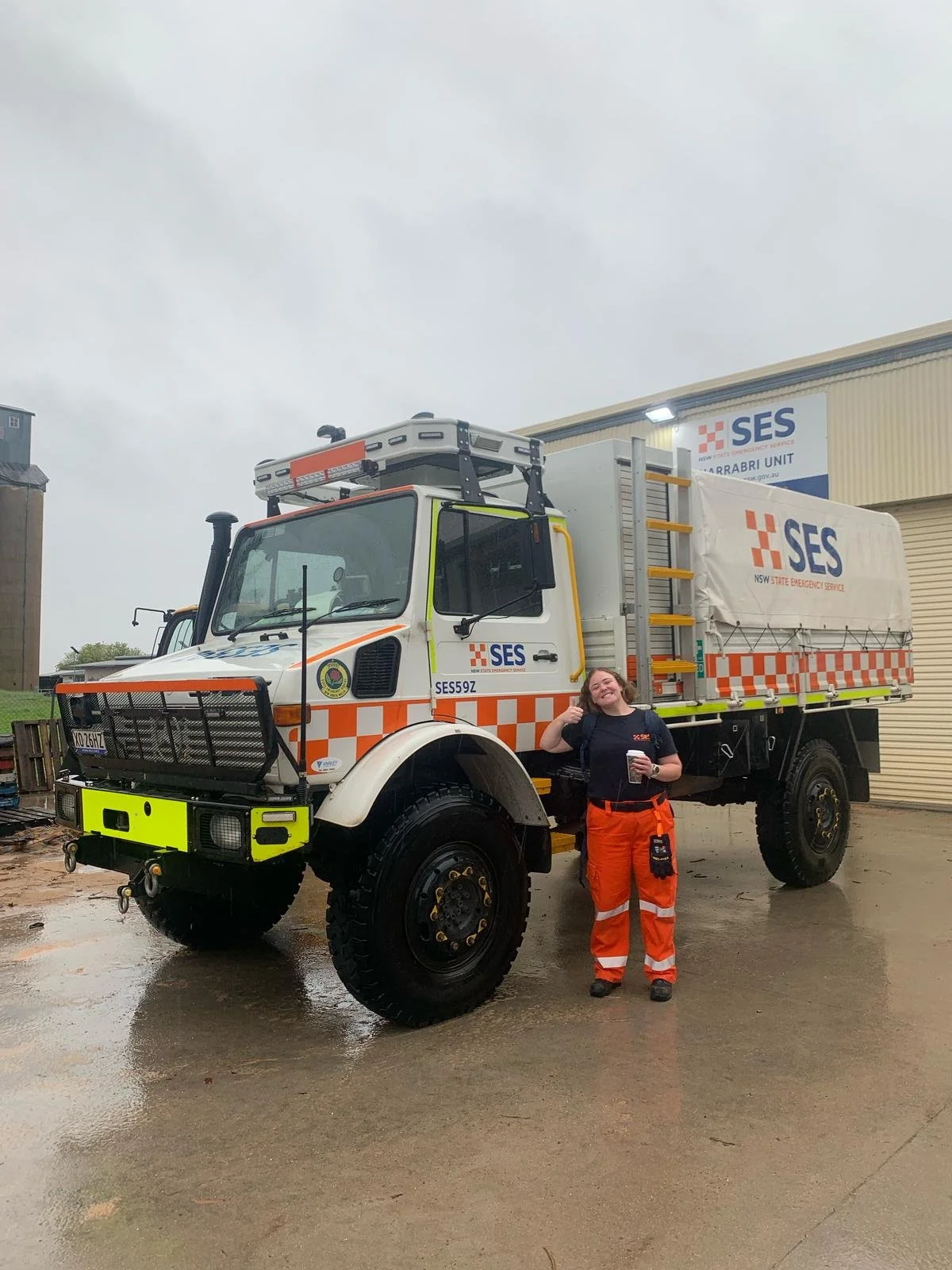 A person standing next to a large emergency service truck, holding a coffee cup, smiling, with an overcast sky in the background.