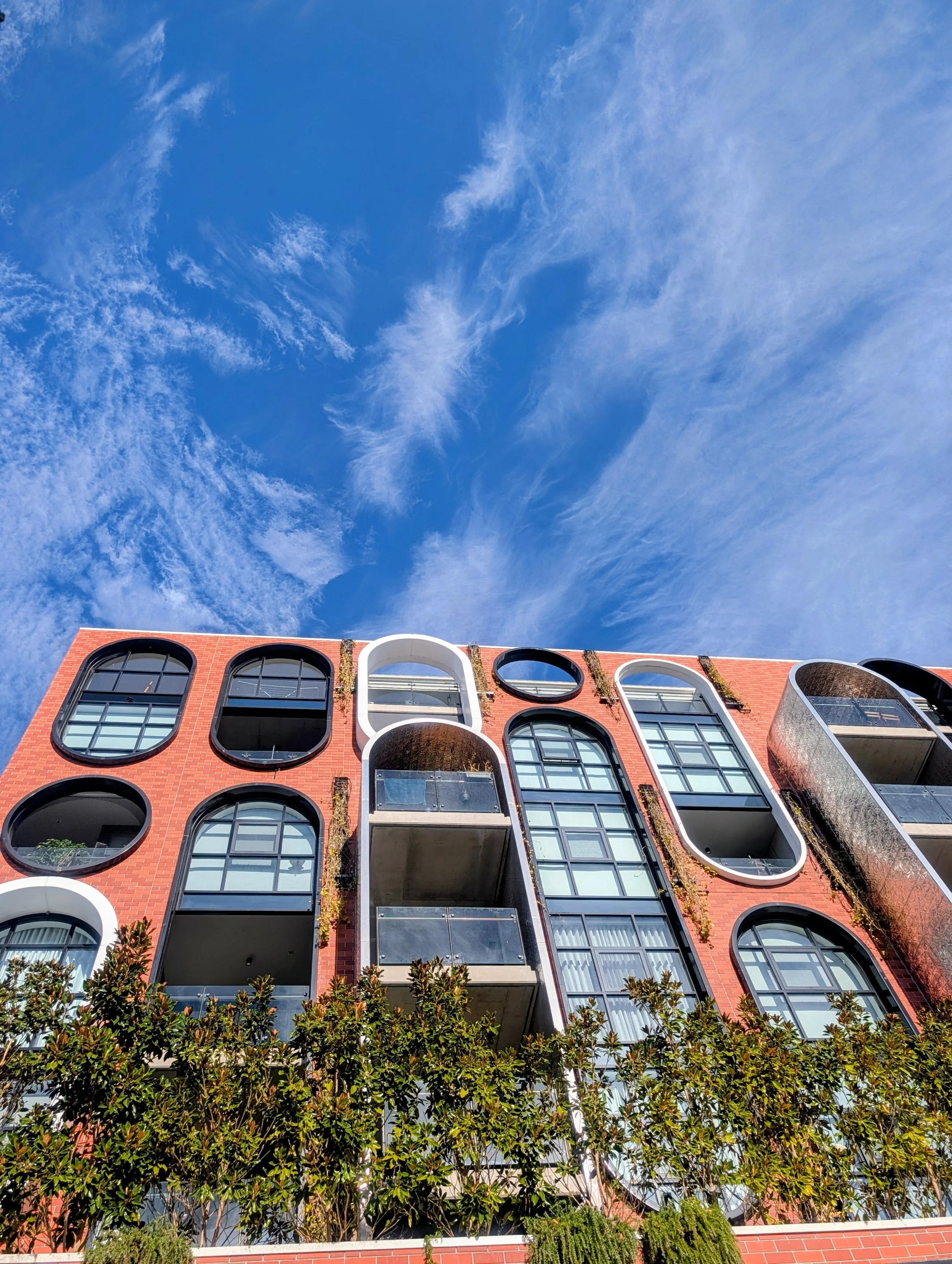 A modern multi-story residential building with a red brick facade and distinctive large, black-framed, irregularly shaped windows. There are balconies with glass railings, some with visible patio furniture, and greenery, including trees and plants, i