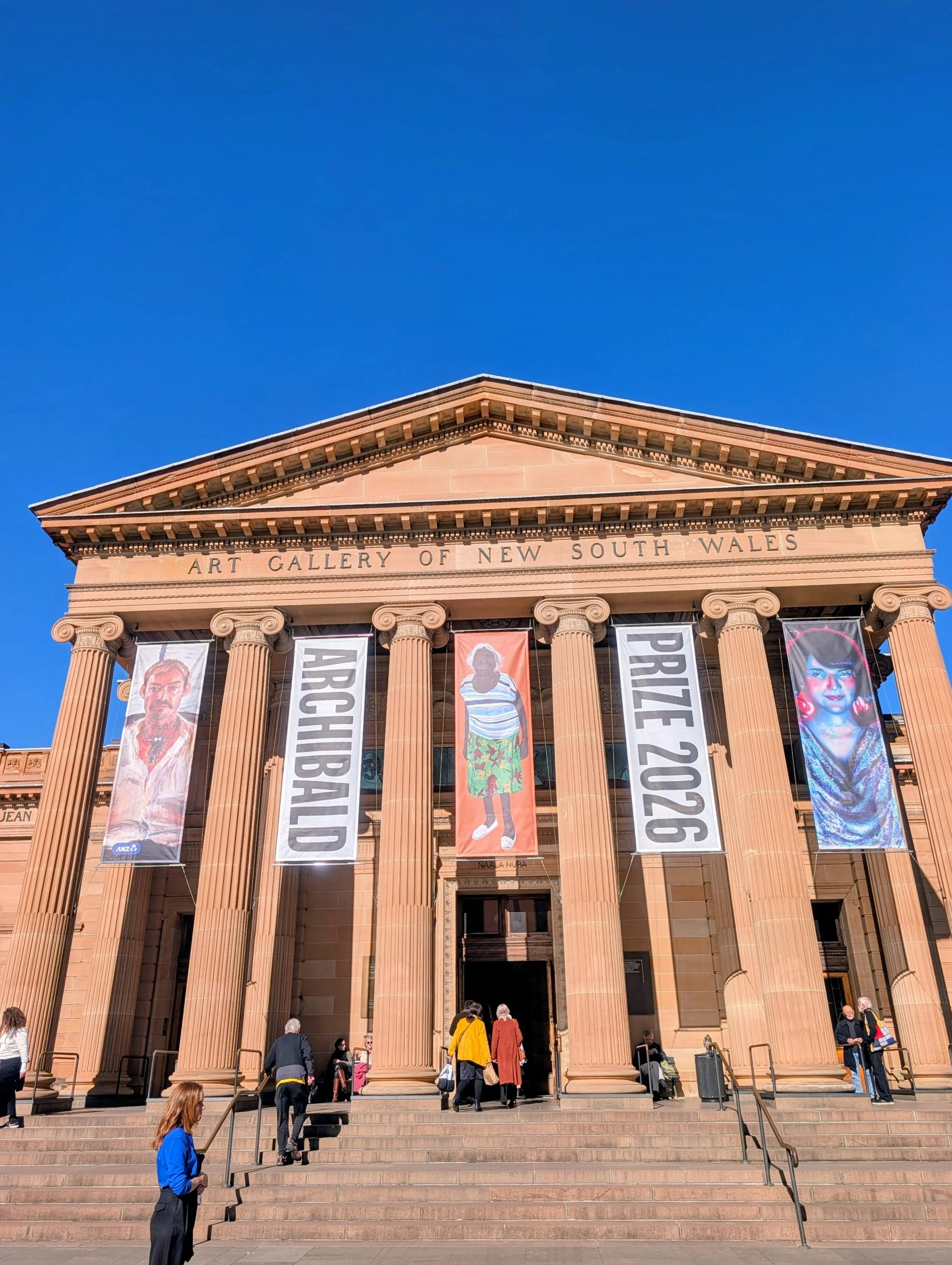 The Art Gallery of New South Wales building with classical columns and banners showcasing artists' portraits, with visitors ascending the steps.