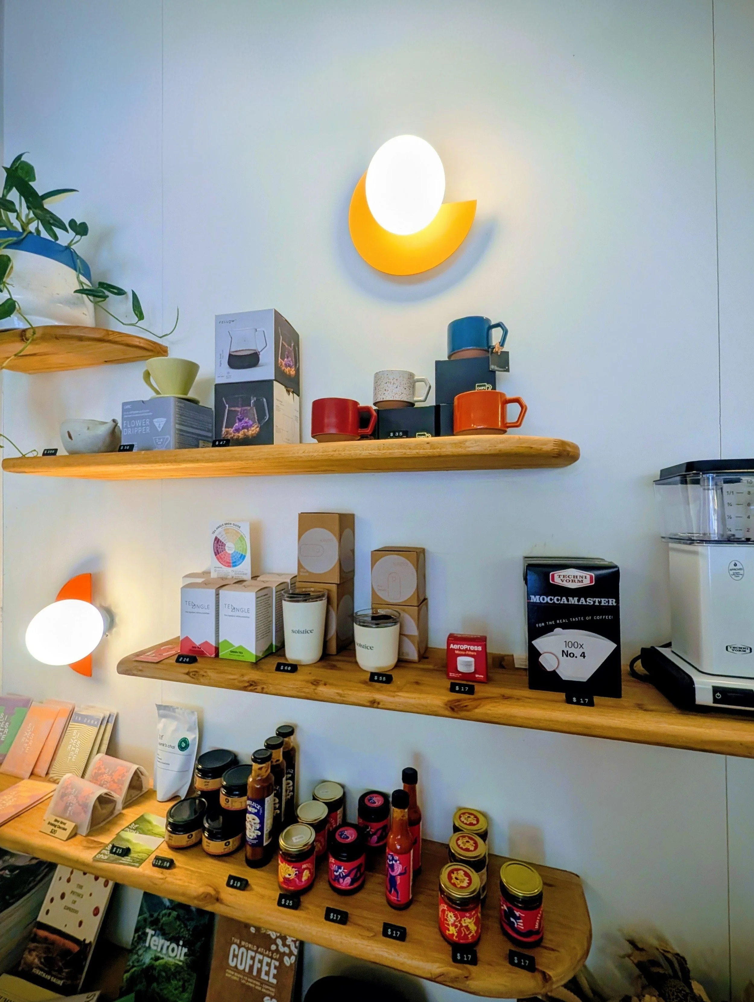Display of coffee mugs, jars, and containers on wooden shelves in a store, with books and product boxes nearby.