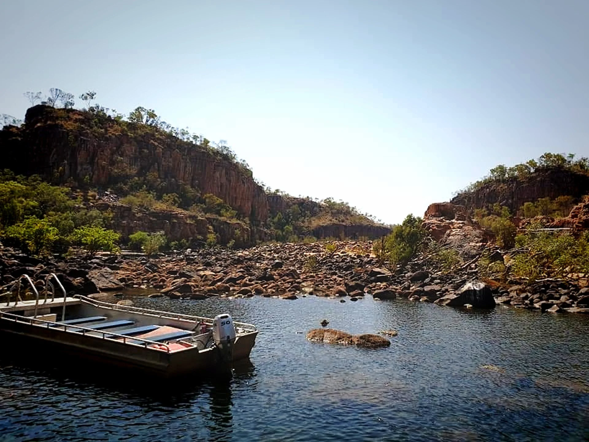 Boat with solar panels on a body of water, surrounded by rocky shores and rugged hills with sparse trees under a clear sky.