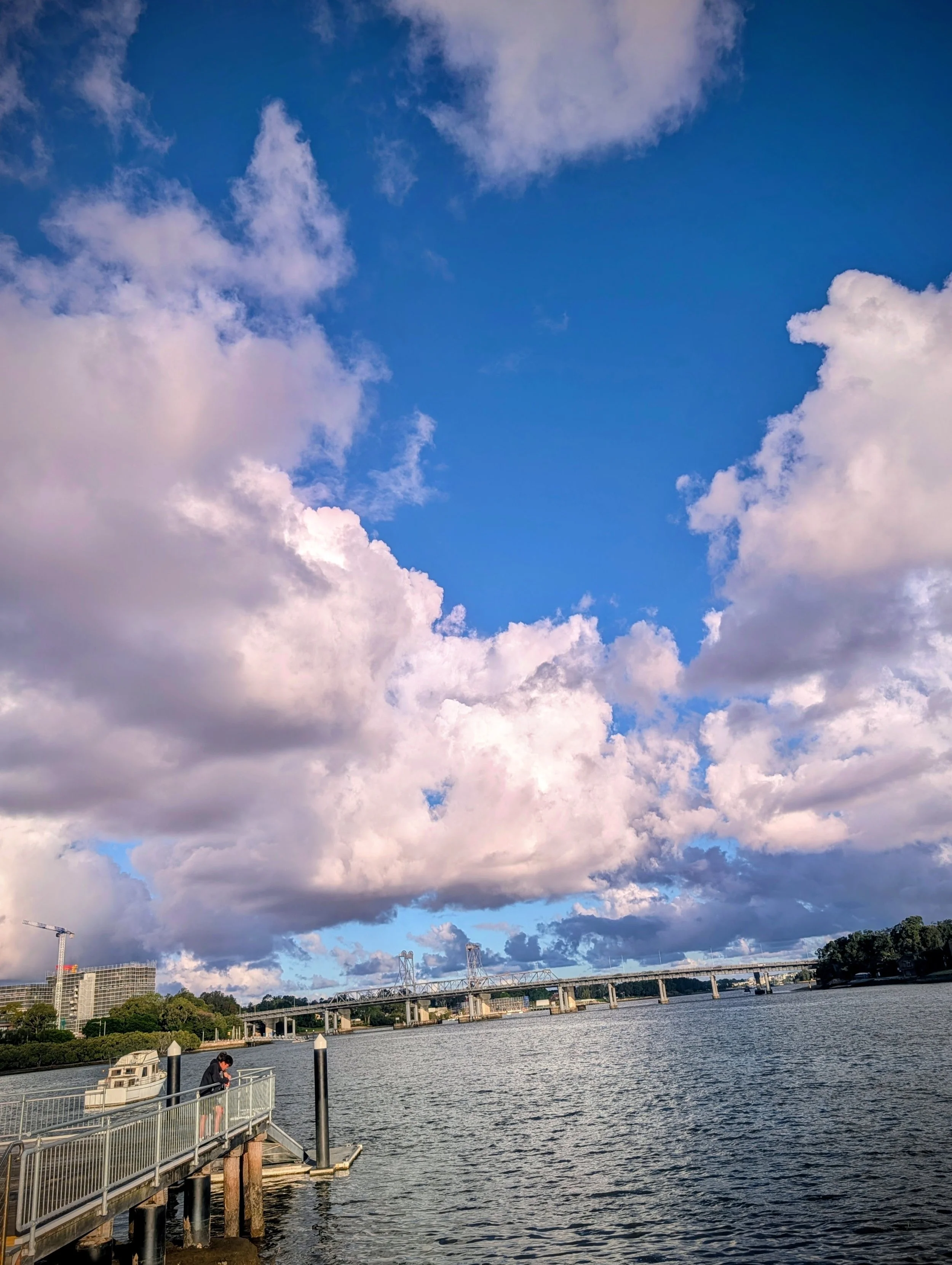 View of a river with a bridge and urban buildings in the background under a sky with scattered clouds.