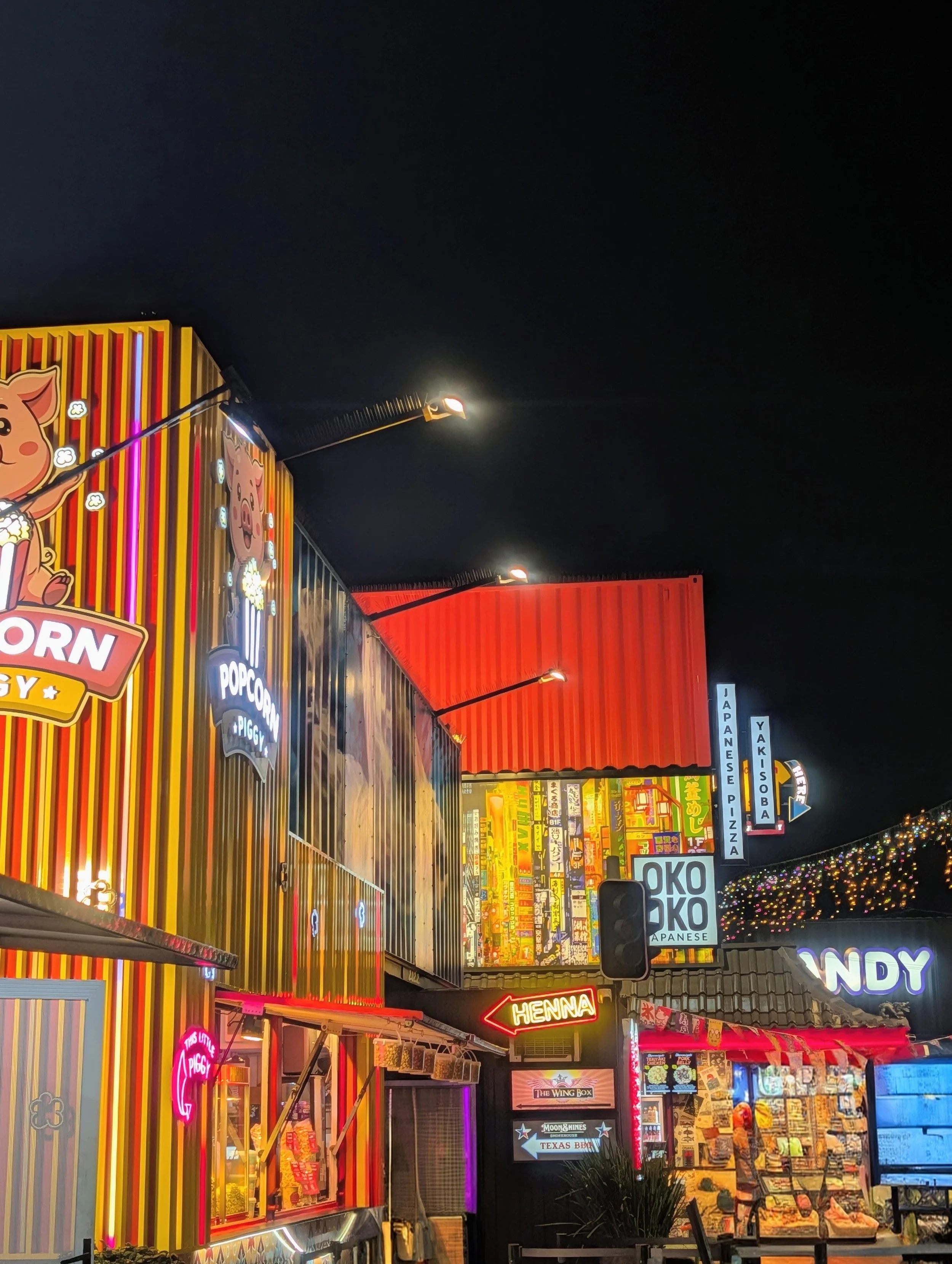 Night scene of a colorful shopping area with illuminated signs and storefronts, including a popcorn stand with a pig mascot, a neon sign for henna, and various shops advertising Japanese cuisine, pizza, and Texan barbecue, with string lights and a dark sky above.