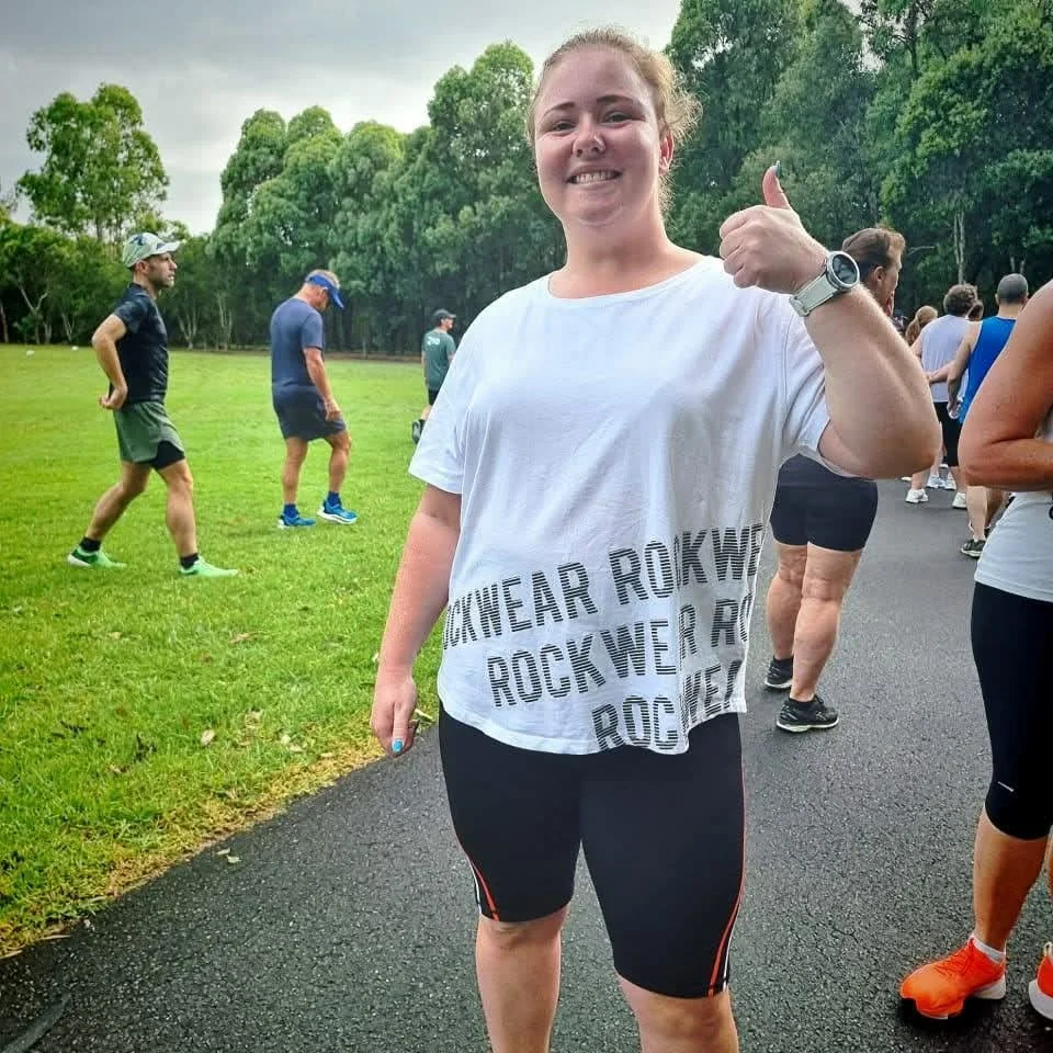 Smiling woman in white T-shirt and black shorts giving a thumbs-up at a group run or race outdoors on a paved path with green grass and trees in the background.