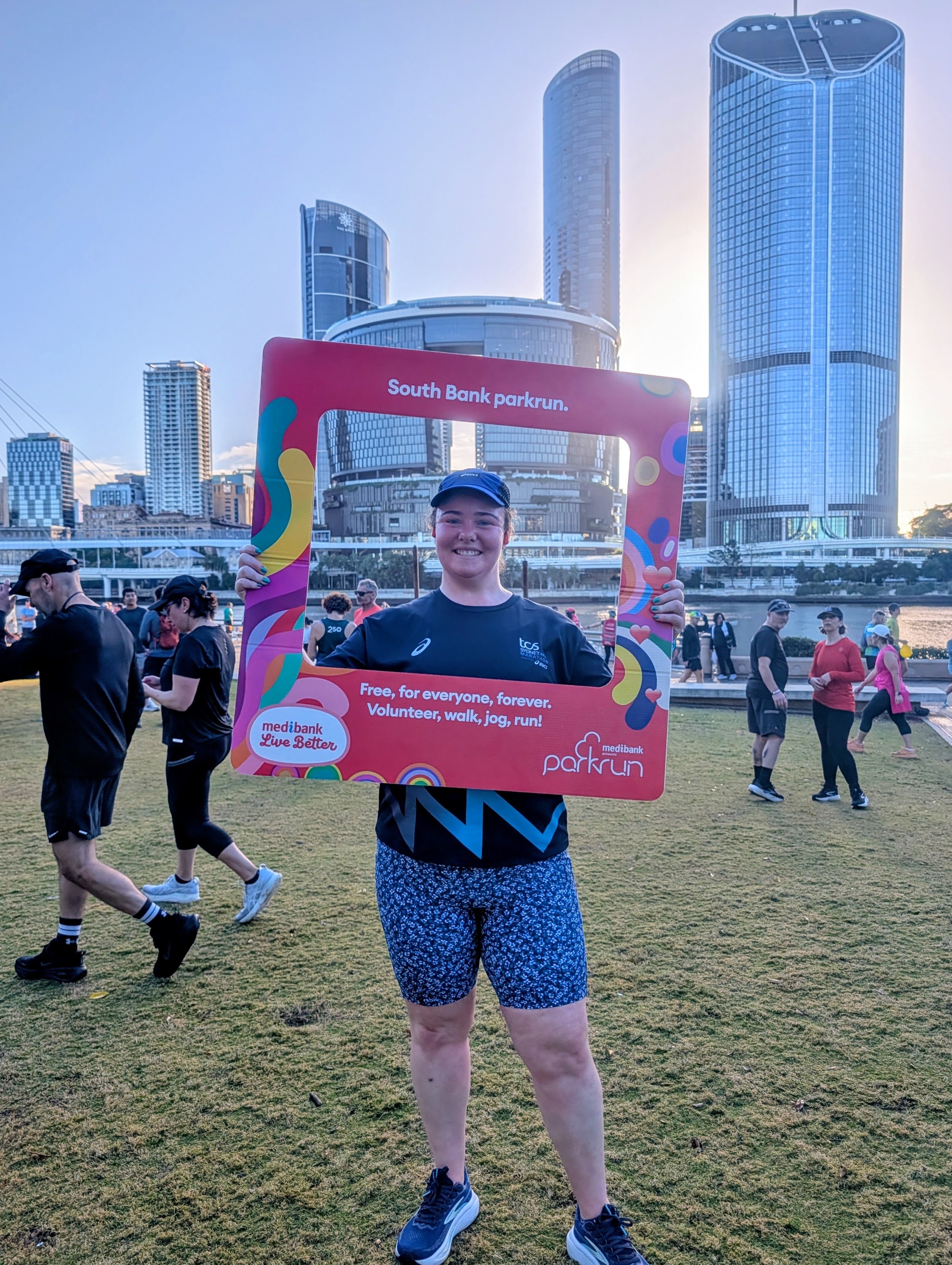 A smiling woman in workout clothes holding a colorful photo frame that reads 'South Bank parkrun' and 'Free for everyone, forever. Volunteer, walk, jog, run!' with a city skyline background and other people in the park.
