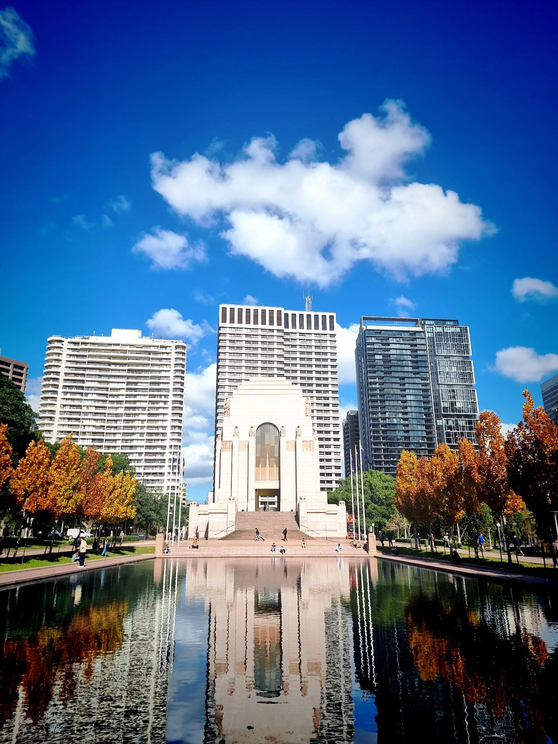 A cityscape featuring tall buildings, a rectangular monument with stairs, trees with orange and green leaves, and a reflecting pond with trees and buildings reflected in the water under a partly cloudy blue sky.