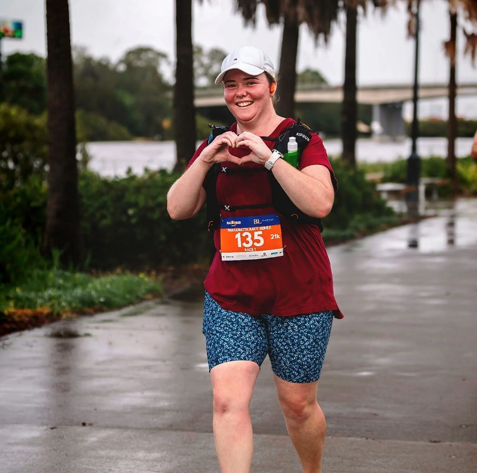 Woman running in a marathon, making a heart with her hands, wearing a white cap, red shirt, and blue shorts, with a race bib numbered 135, on a wet pavement with trees and water in the background.