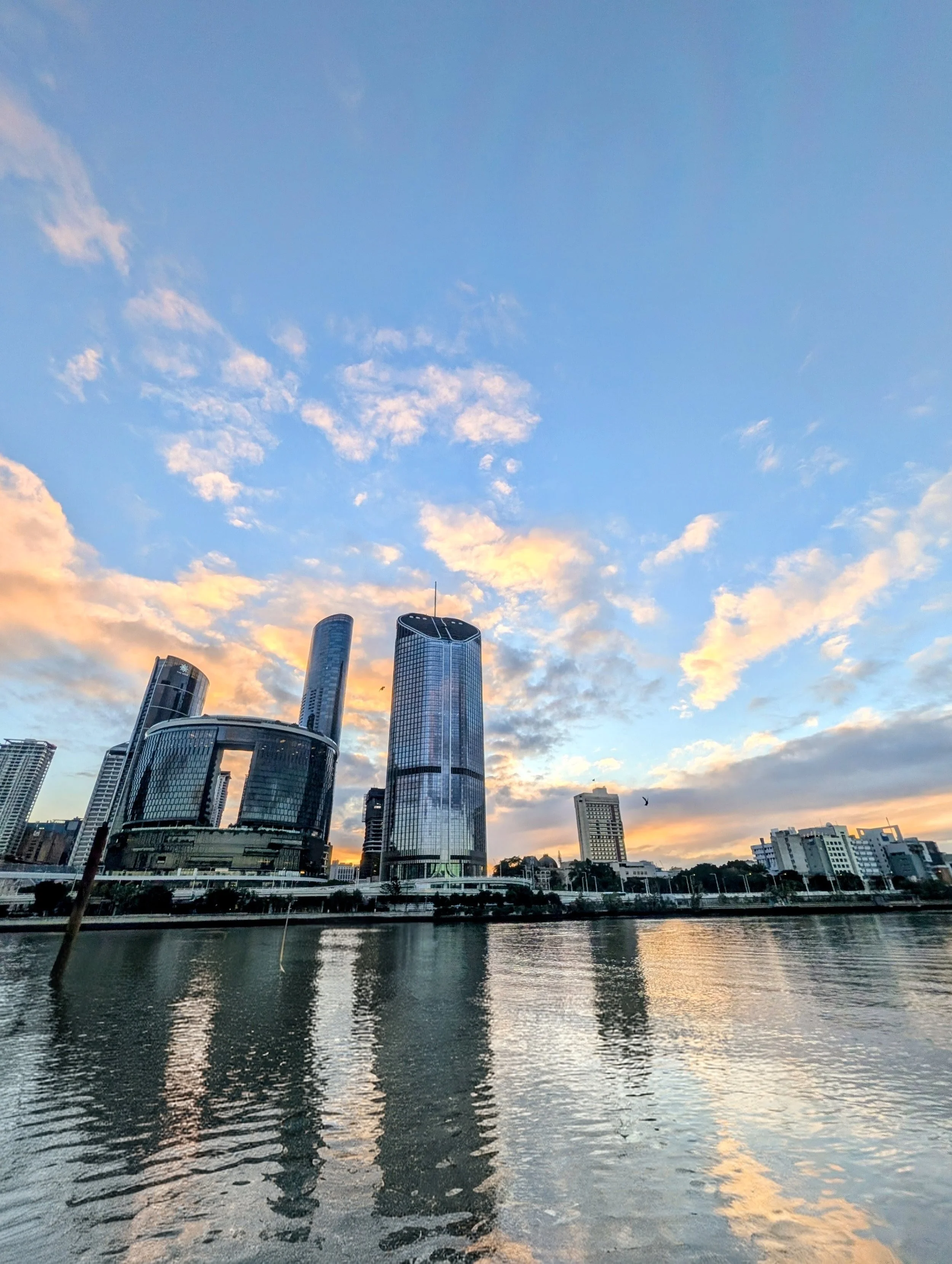 City skyline with modern skyscrapers reflected in a river at sunset, with a partly cloudy sky.