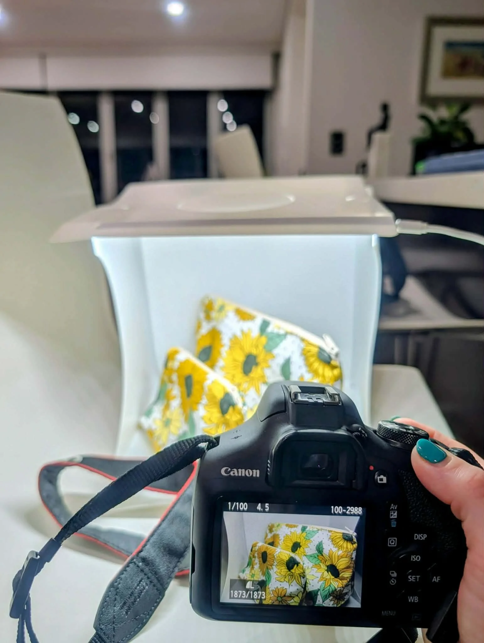 Photo of a camera screen capturing a floral tissue box with a yellow sunflower design, a person holding the camera with painted nails, a white chair, and a floral bag in the background.