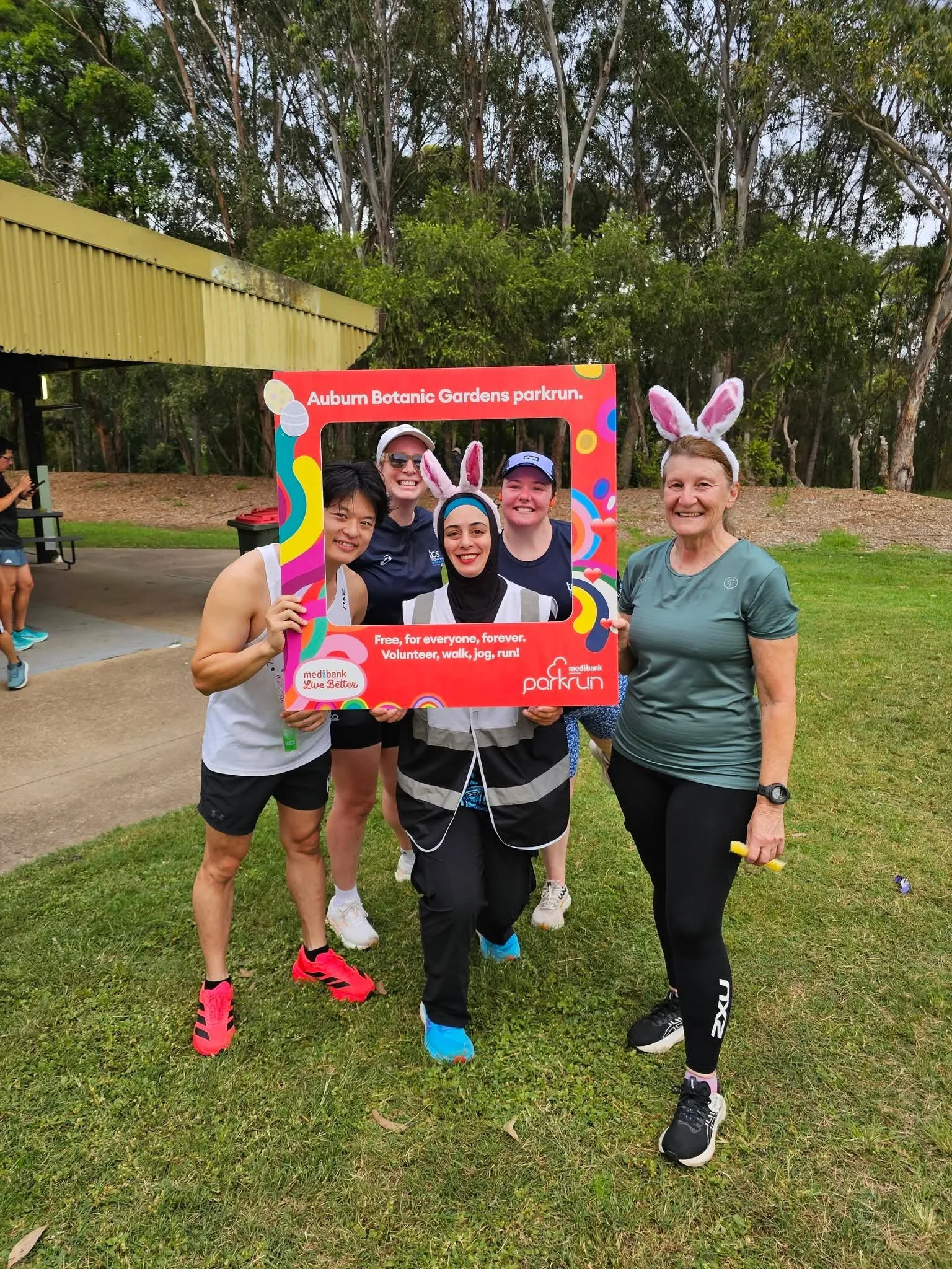 Group of five women, some wearing bunny ears, standing outdoors on grass, posing with a colorful frame at Auburn Botanic Gardens park run event.