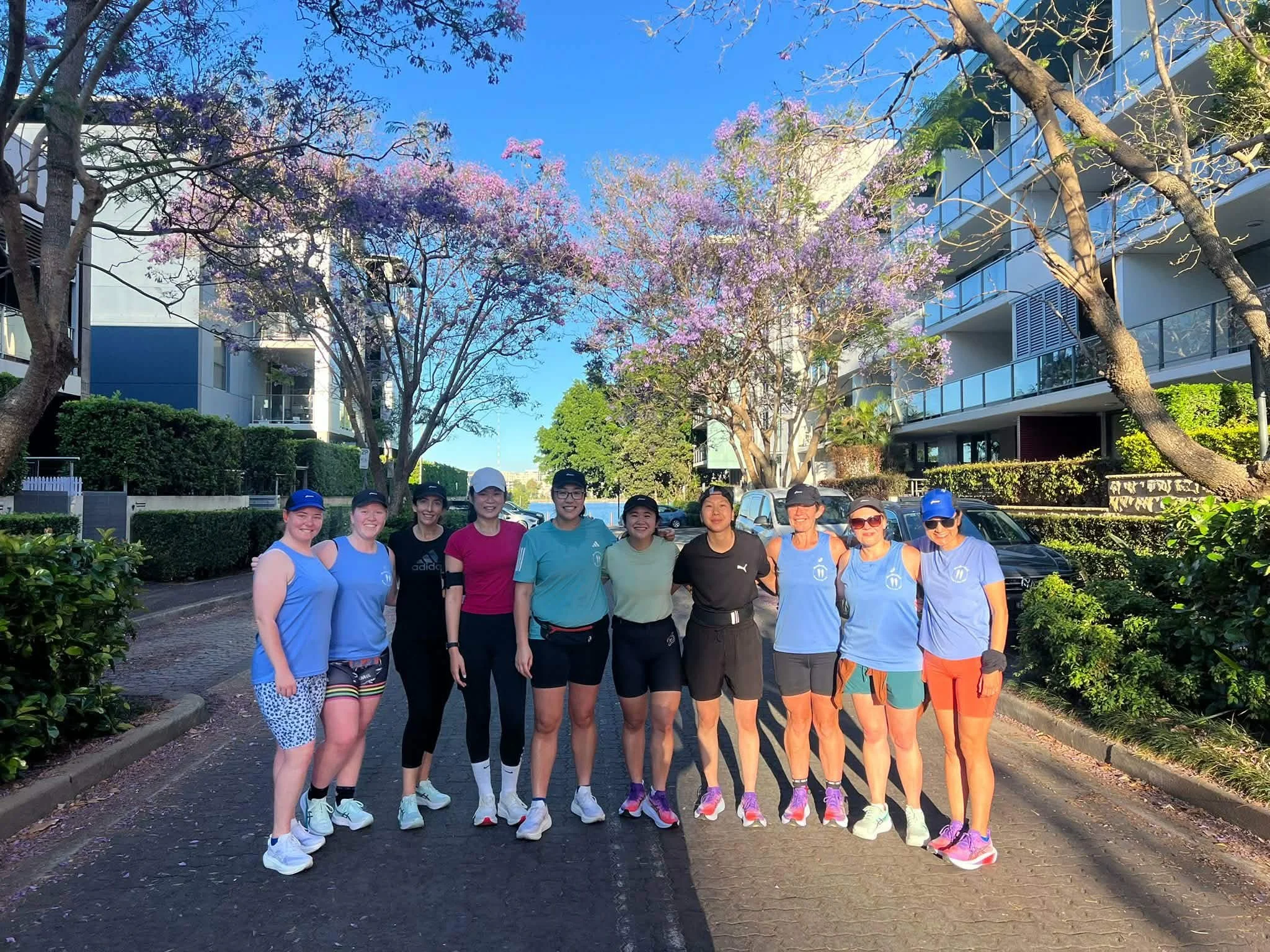 Group of women runners standing outdoors on a sunny day, with trees and apartment buildings in the background.