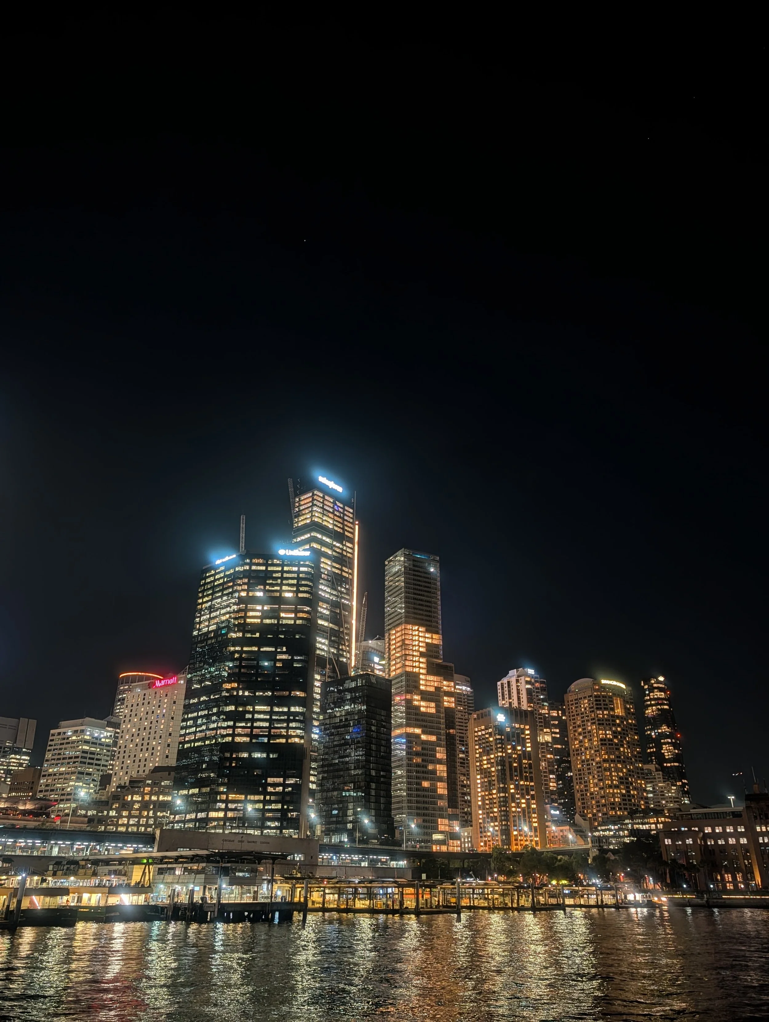 Nighttime skyline of a city with illuminated skyscrapers reflected in a body of water in the foreground.