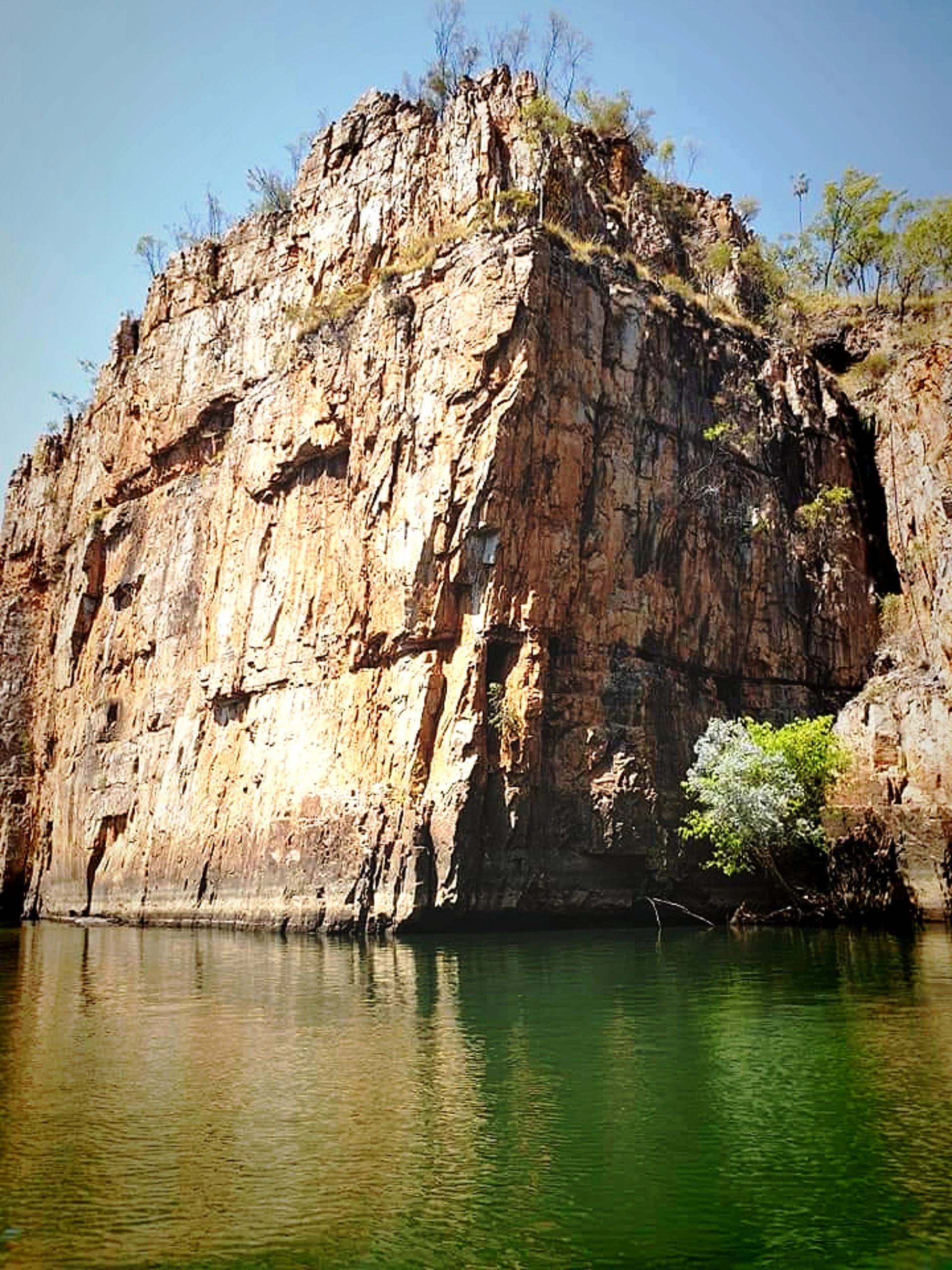 A large, rocky cliff with sparse vegetation is reflected in a calm river below. The sky above is clear and blue.