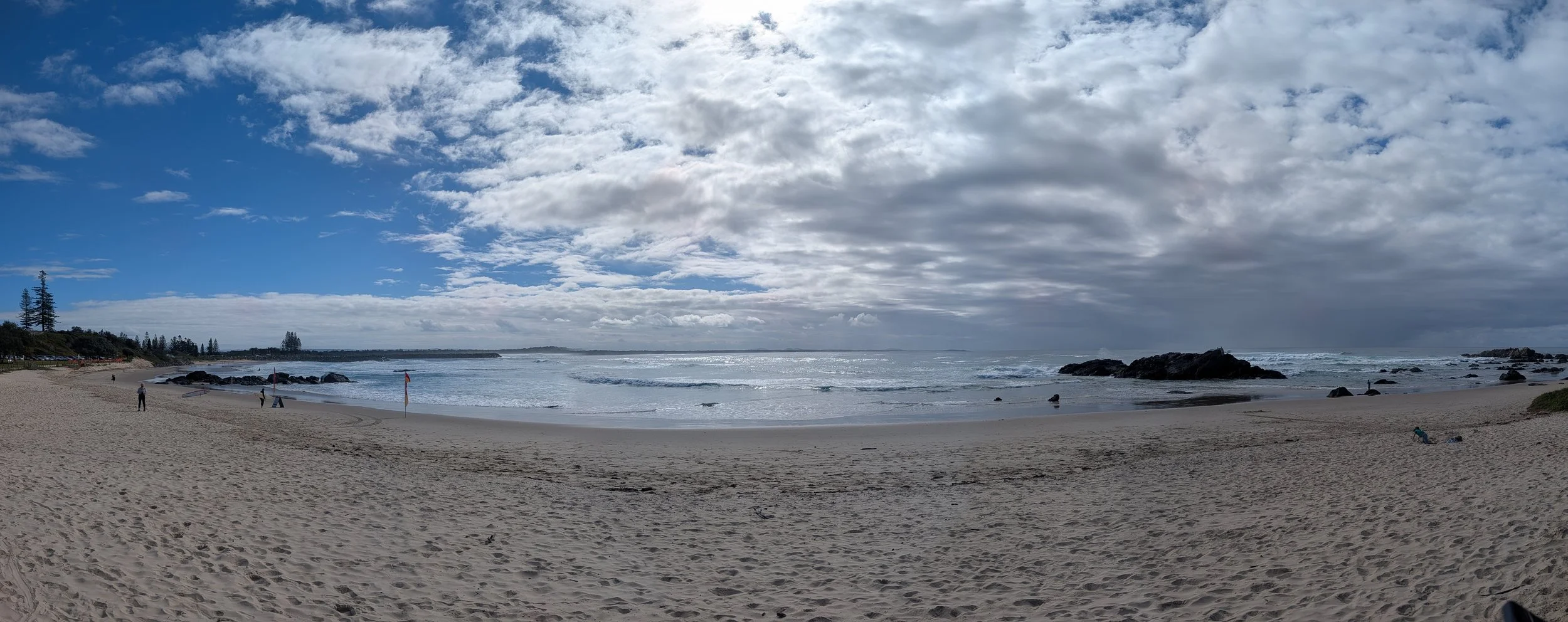 A beach with sandy shoreline, rocky outcroppings, and a few people. The sky is partly cloudy with blue patches and clouds overhead, and the ocean waves gently crash onto the shore.