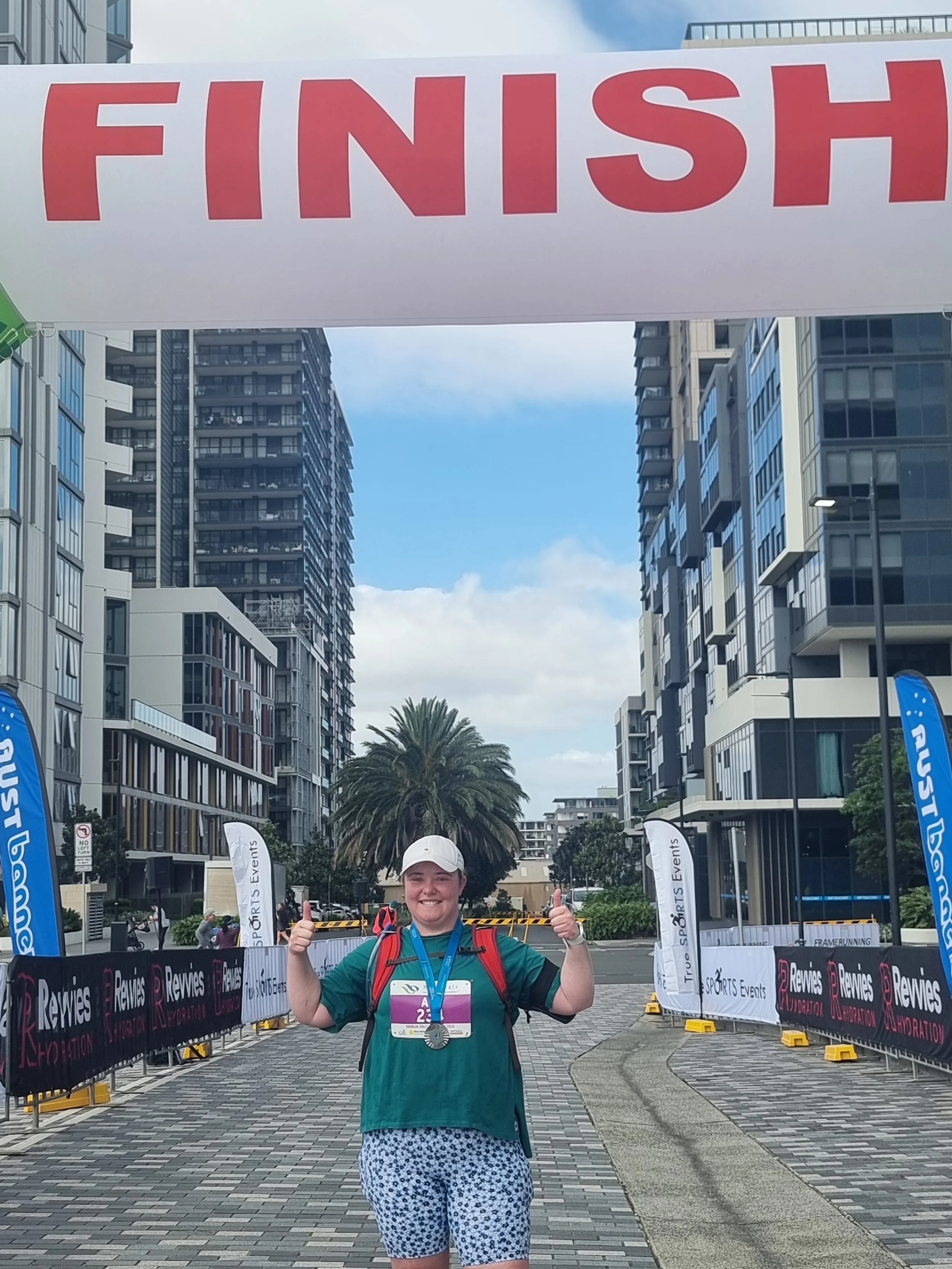 A smiling woman with a white cap, green shirt, and patterned shorts stands under a finish line banner at an outdoor event, giving a thumbs-up with both hands. She wears a medal around her neck and an event bib on her chest.
