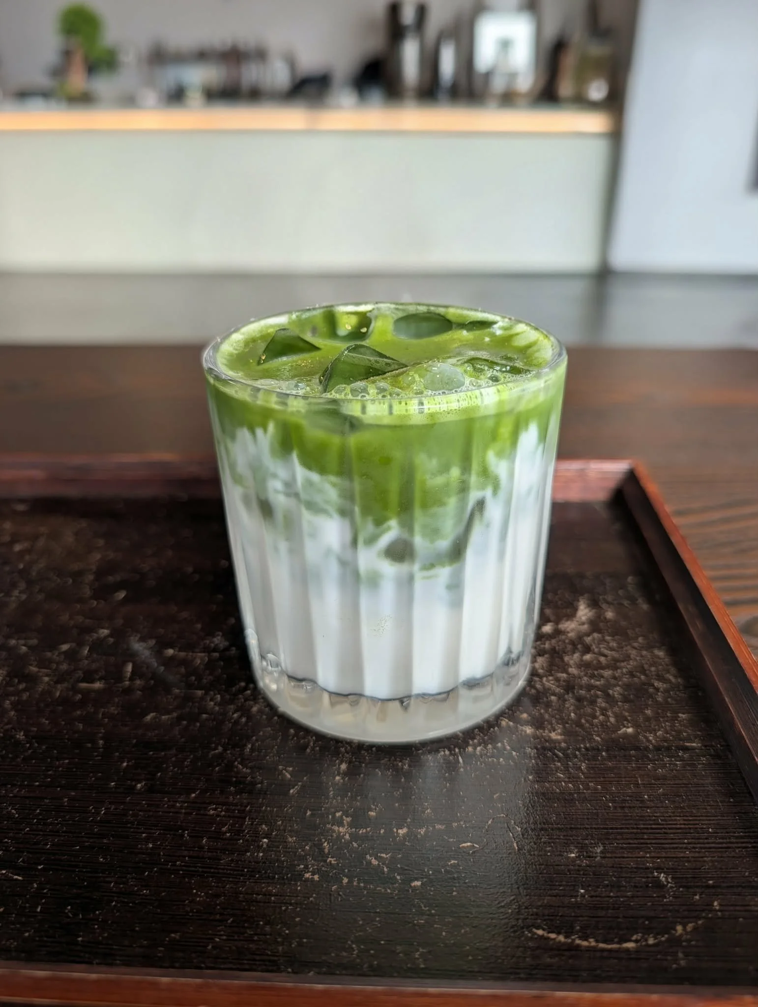 A glass of green matcha latte with ice cubes on a wooden table in a cafe setting.
