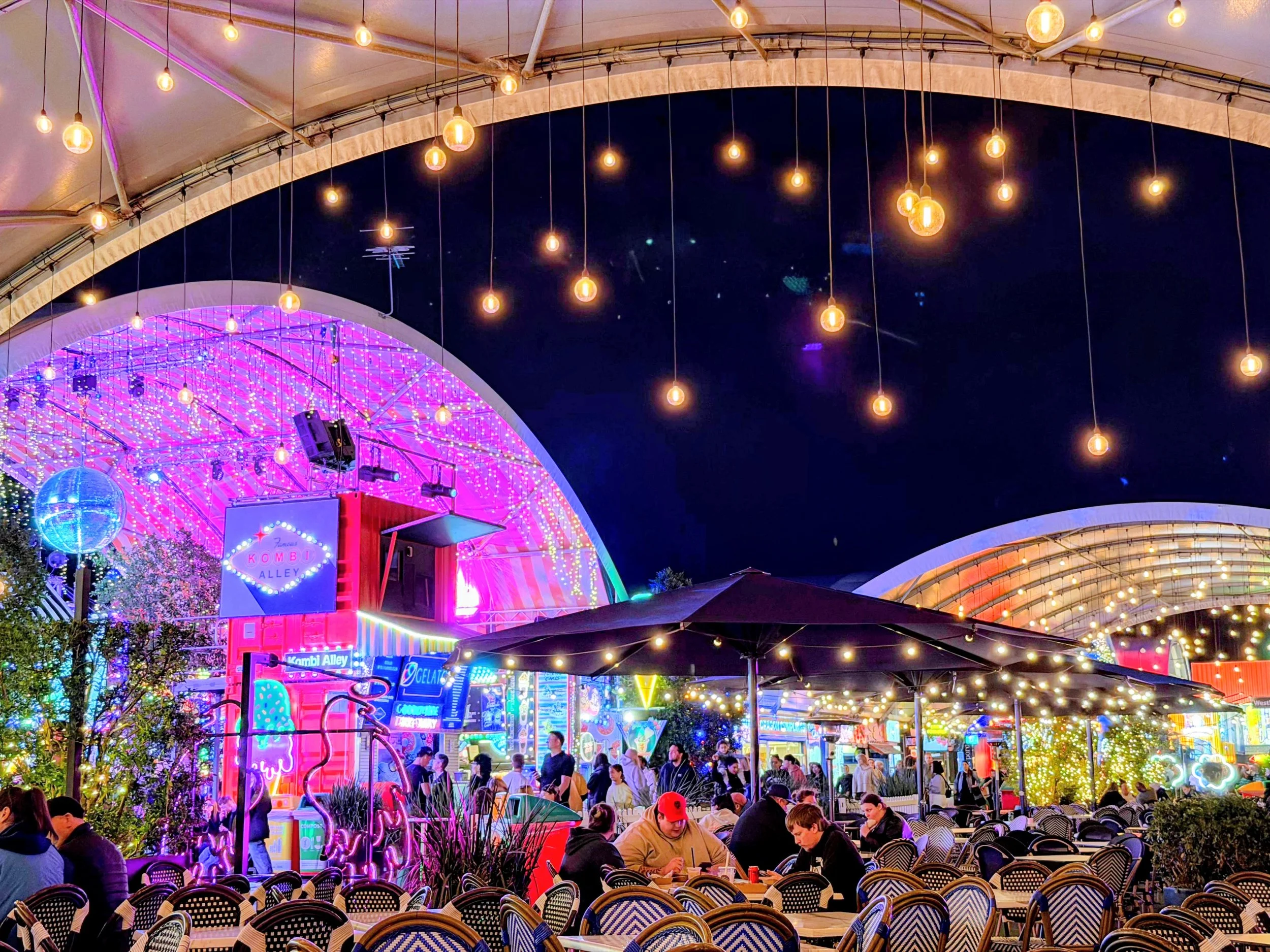 Outdoor amusement park at night with colorful neon lights, hanging string lights, and many people sitting and walking around.
