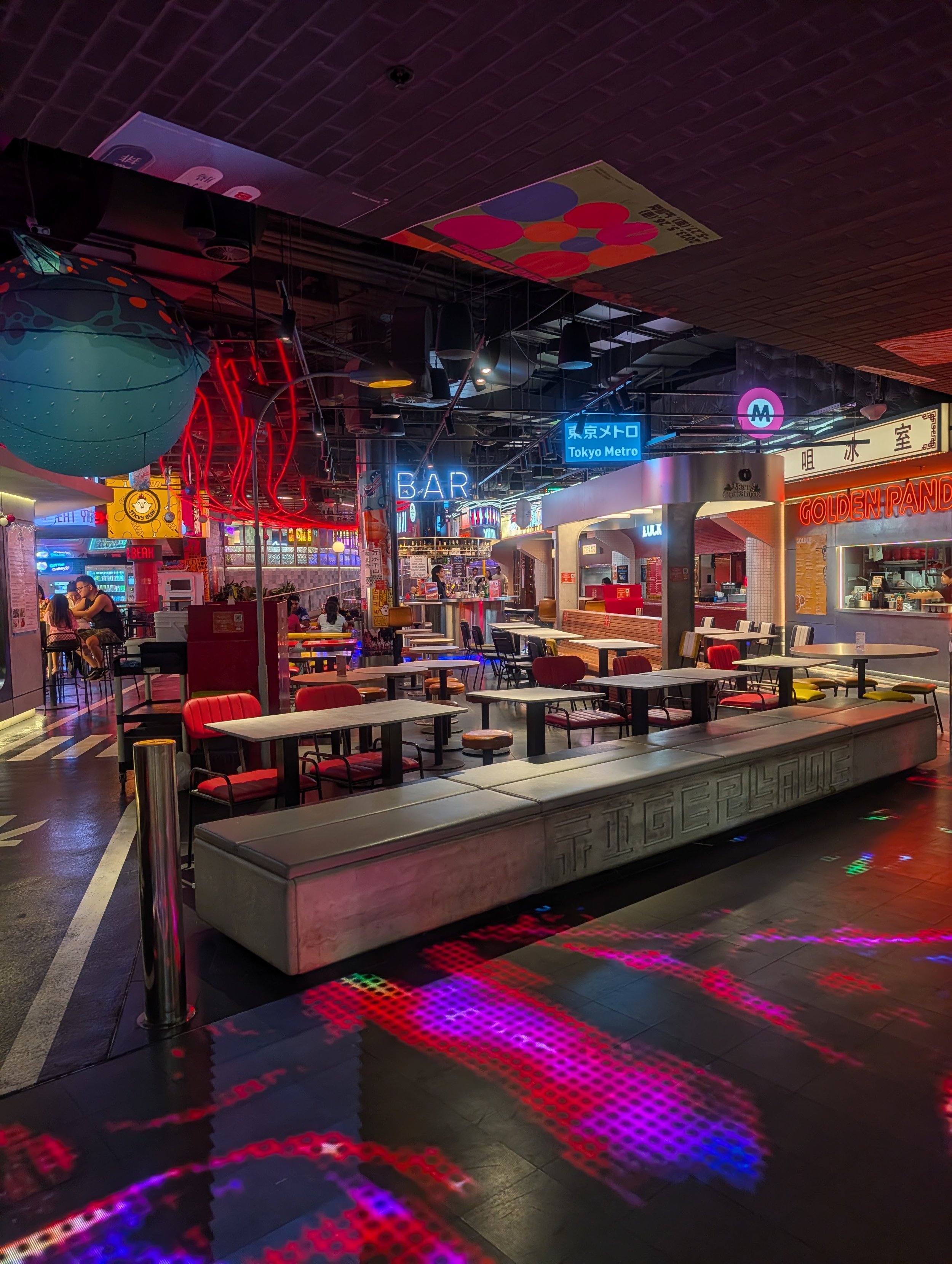 Interior of a modern entertainment or dining venue with colorful neon signs, tables, chairs, and decorative lighting.