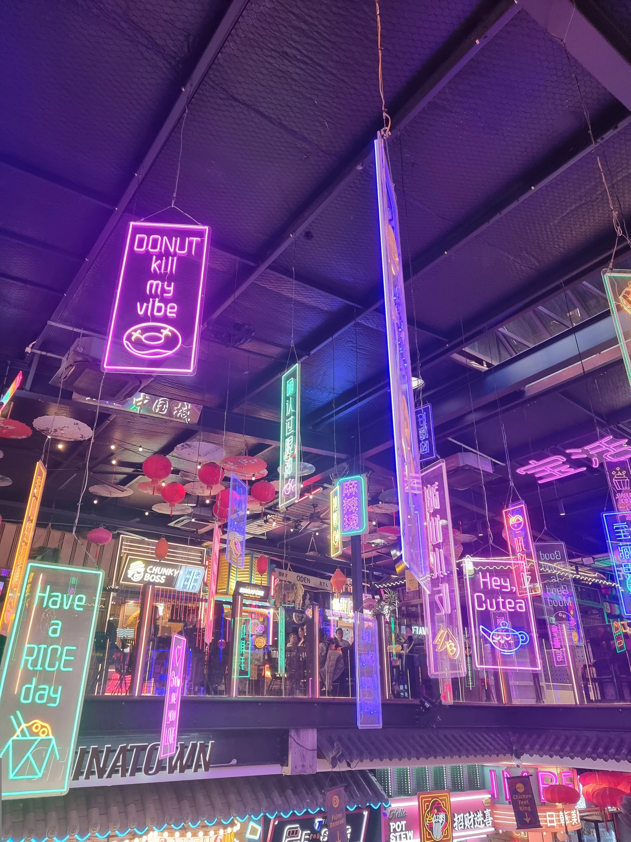 Nighttime scene of a shopping or entertainment area with colorful neon signs displaying various messages, some in English and others in Chinese, with hanging red lanterns and reflections on glass surfaces.