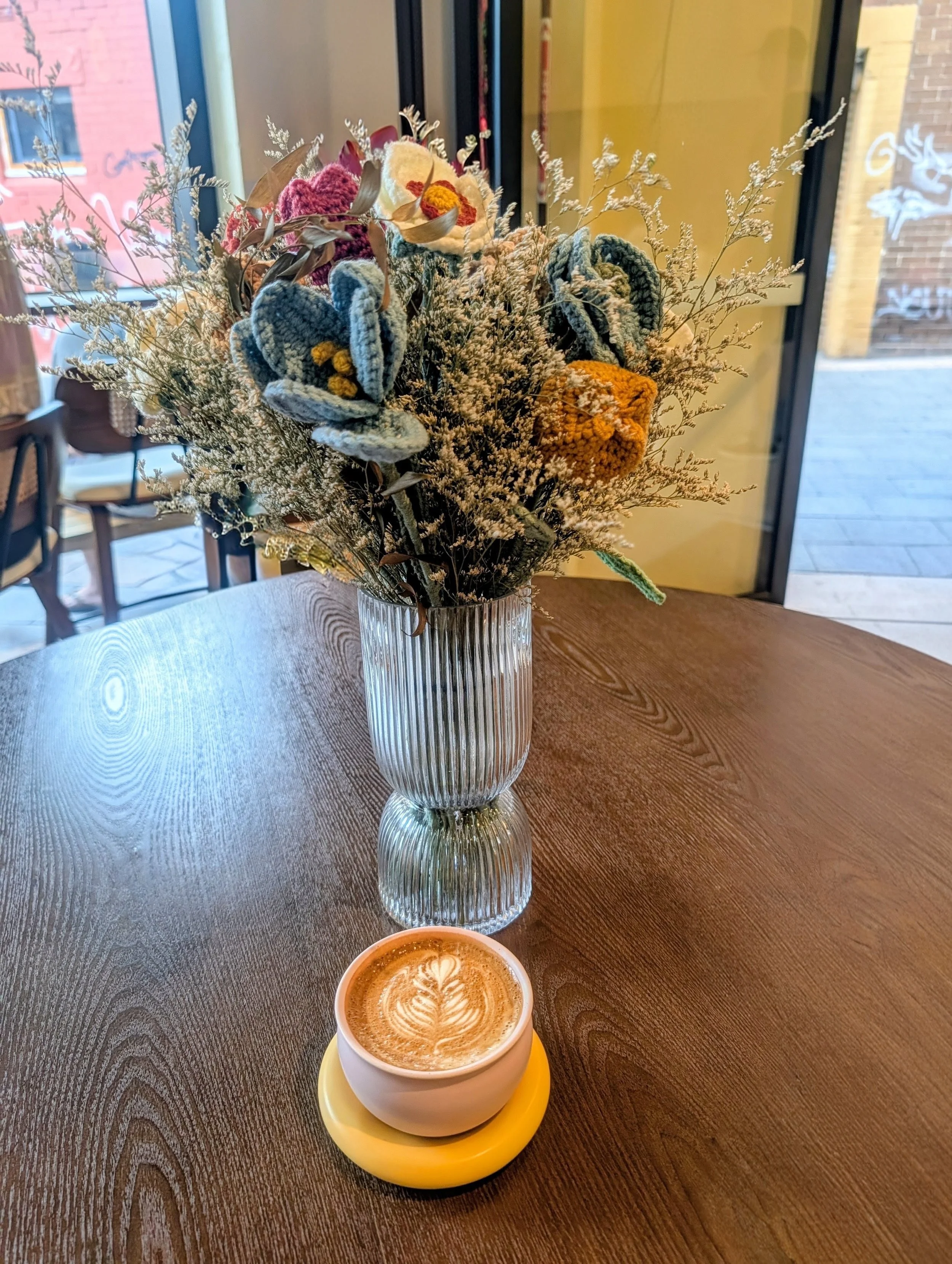 A bouquet of colorful dried flowers in a tall, textured glass vase on a wooden table, with a cup of latte featuring latte art in the foreground.