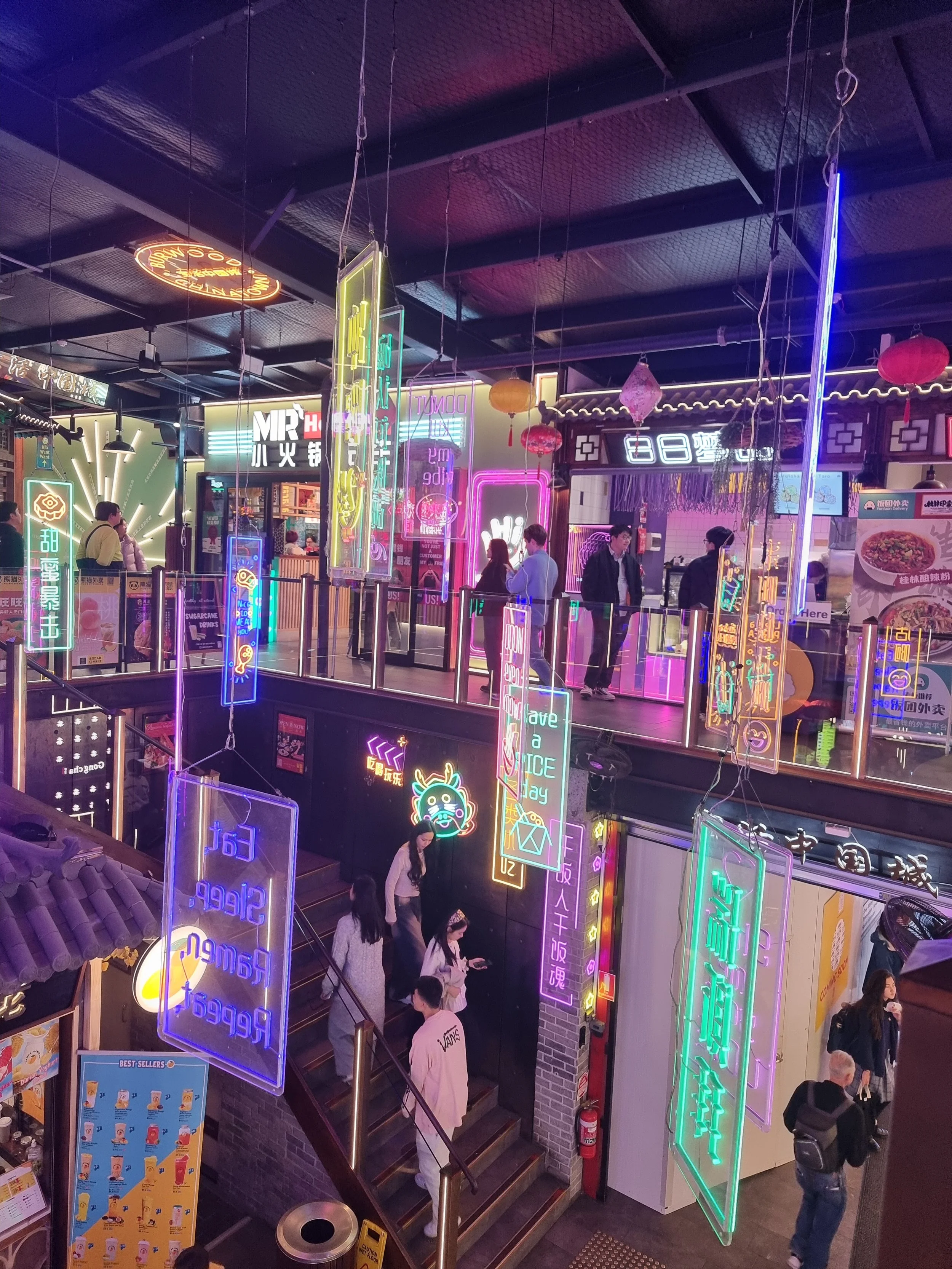 Interior of a shopping mall or marketplace with neon signboards in various languages, people walking, and a staircase.