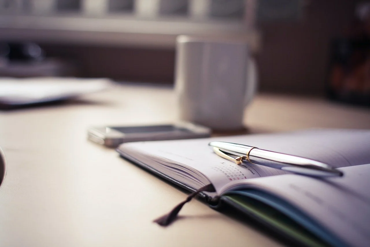 Desk with open planner, pen, smartphone, mug, and blurred background.