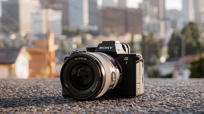 A Sony Alpha 7 mirrorless camera with a zoom lens placed on a rough surface outdoors, with a blurred cityscape in the background.