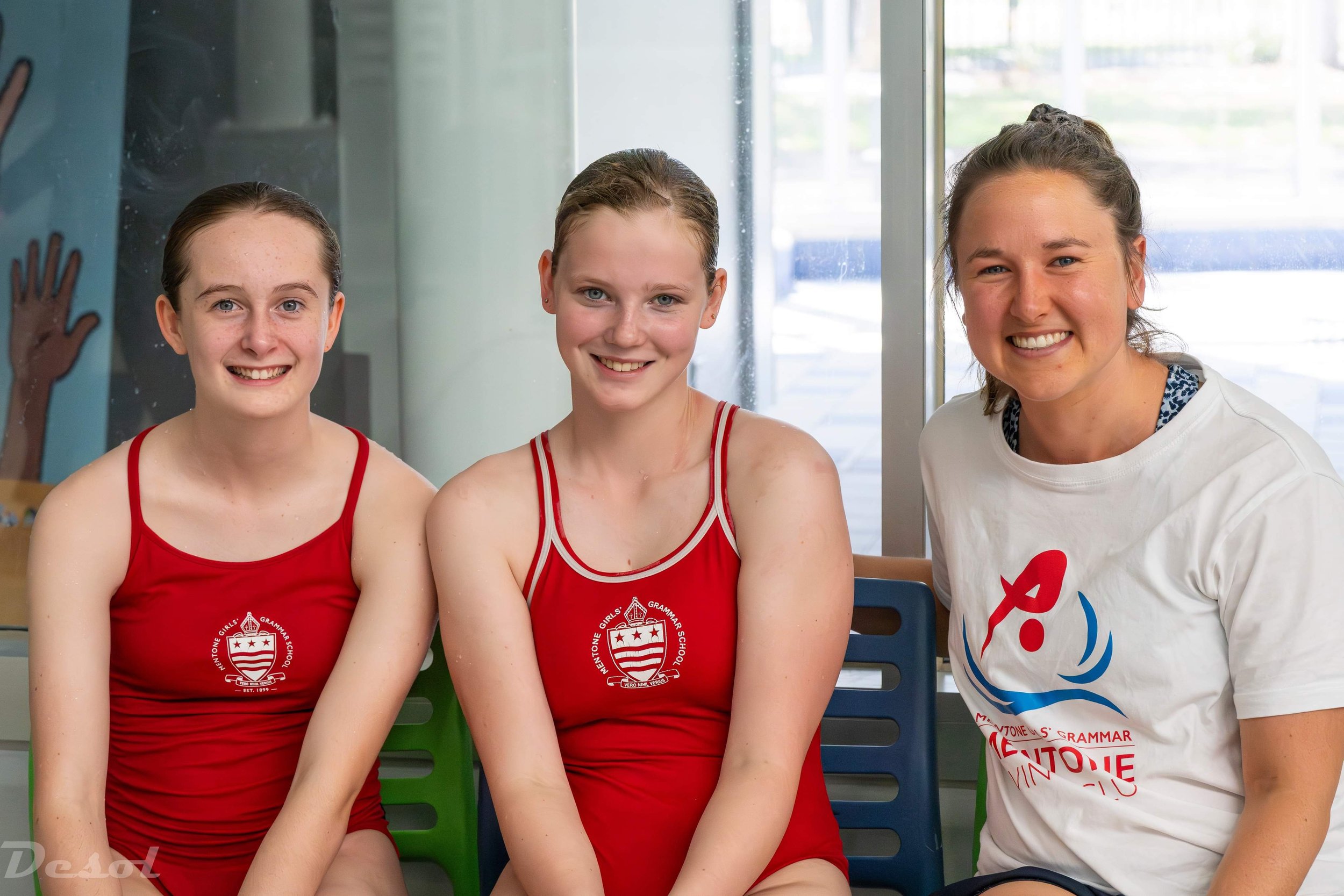 Three young girls, wearing red athletic jerseys with a school logo, and a woman, wearing a white T-shirt with a colorful design, sitting indoors near a glass window, smiling for the photo.