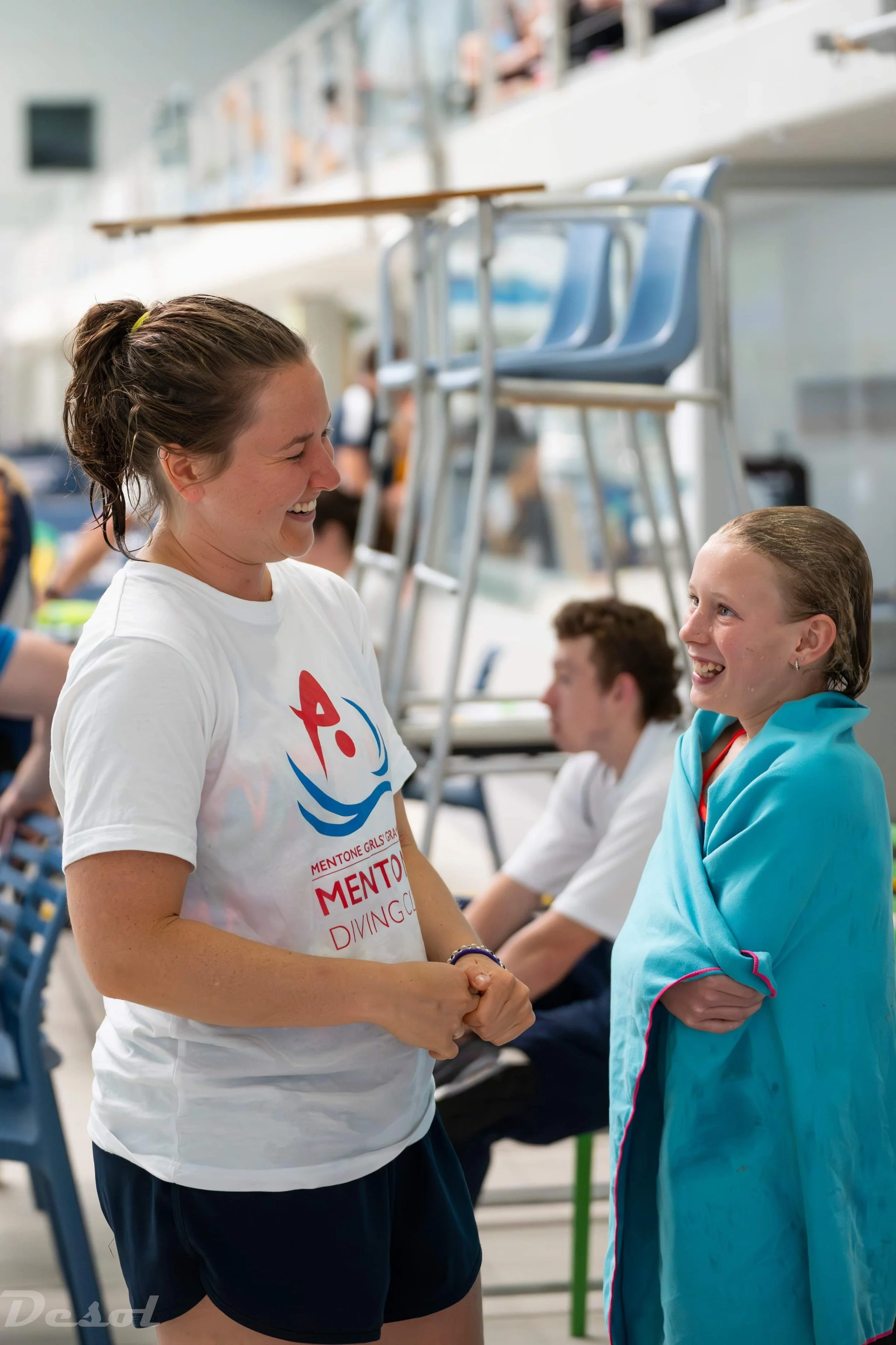A woman and a young girl are smiling and shaking hands at an indoor event. The woman wears a white T-shirt with a logo and text, while the girl wears a blue jacket. They are surrounded by others in the background, seated and standing.