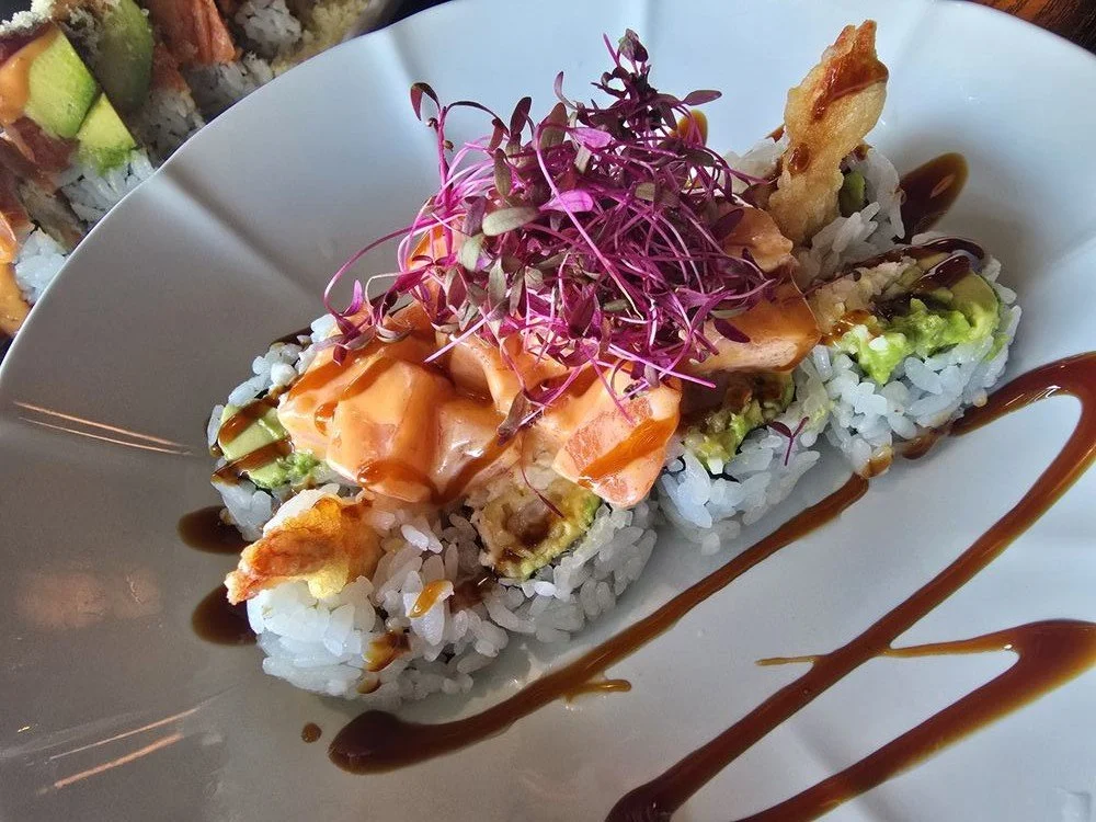 Close-up of a sushi roll topped with microgreens, drizzled with eel sauce, served on a white plate with additional eel sauce drizzled around it.