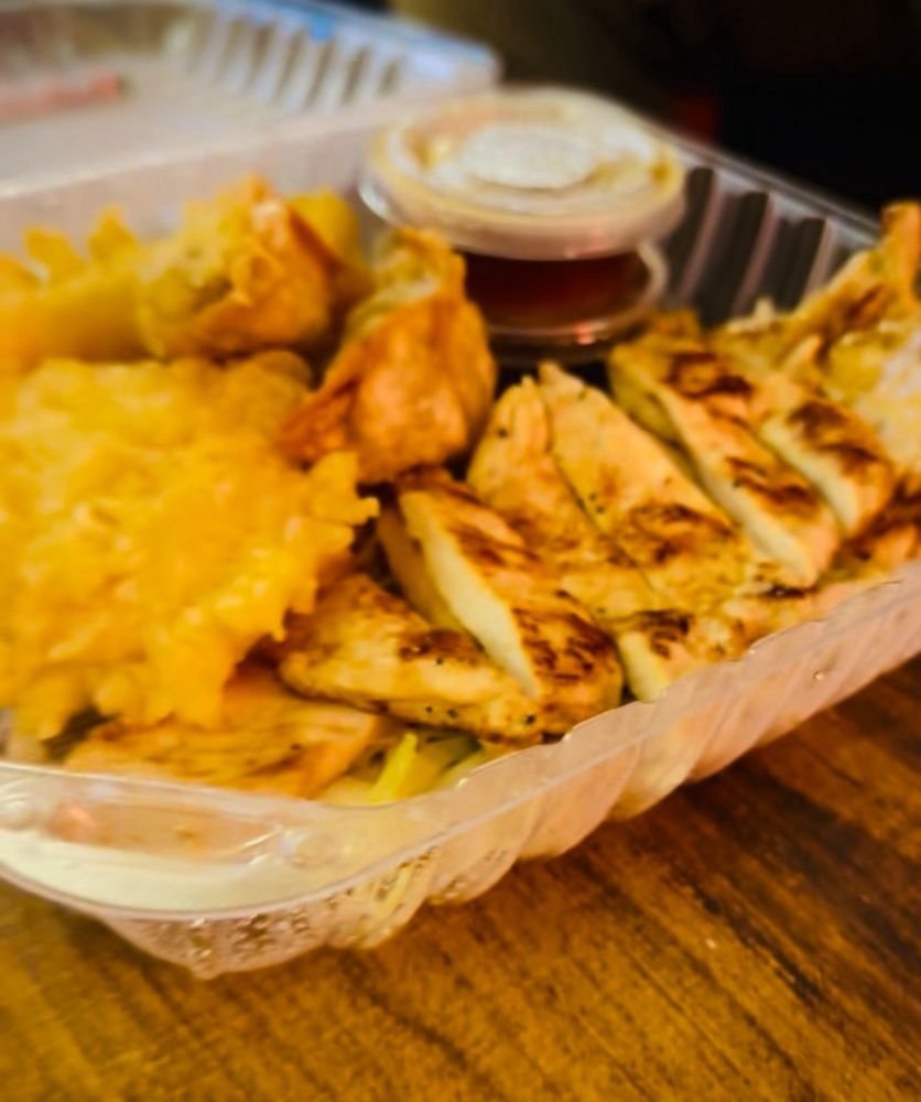 A plastic basket containing fried chicken strips, macaroni and cheese, potato wedges, and a side of dipping sauce on a wooden table.
