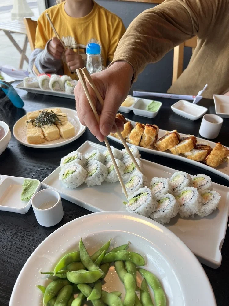 A table filled with various sushi dishes, including rolls, edamame, and sides, with a person using chopsticks to pick up sushi.