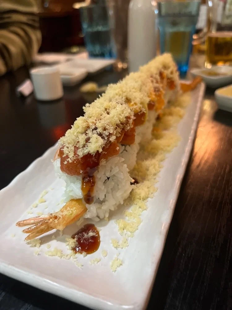 Ebi tempura sushi roll topped with tempura flakes and drizzled eel sauce on a white rectangular plate.