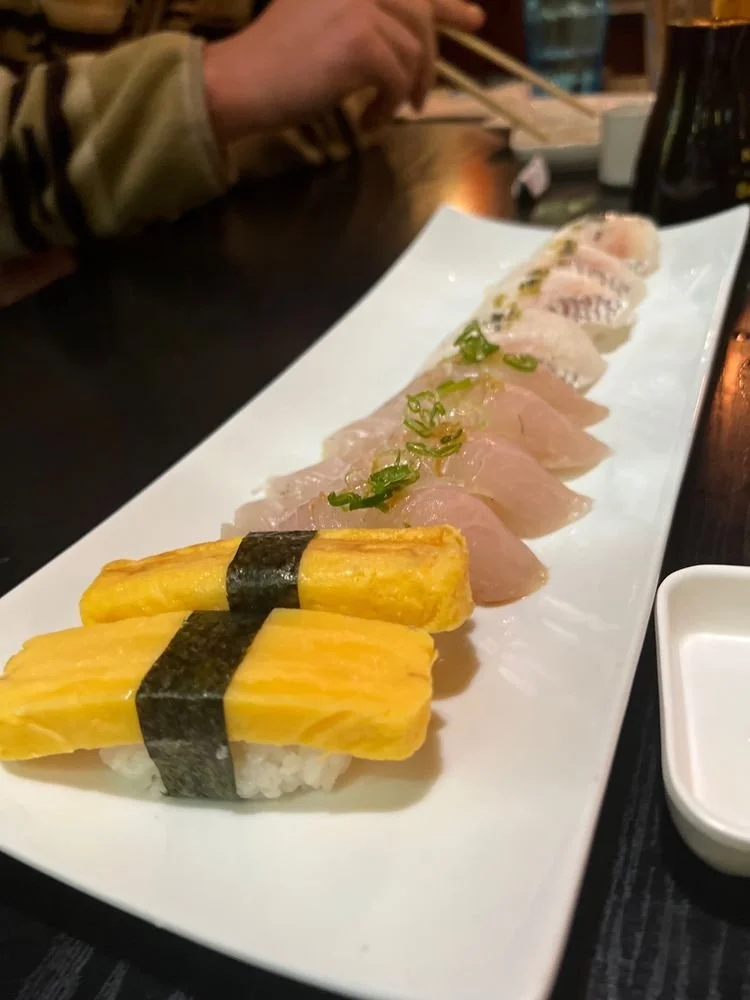 A rectangular white platter with various pieces of sushi, including tamago nigiri in the front, followed by slices of raw fish topped with green onions, and other sashimi pieces in the background.
