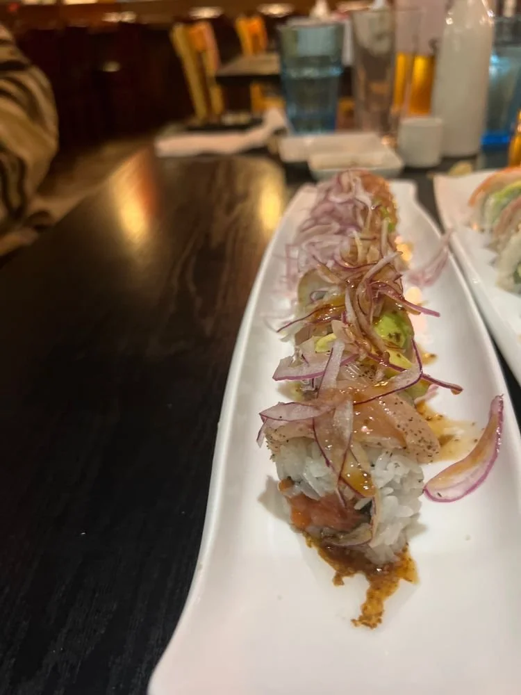 Sushi roll topped with sliced onions and sauce, served on a white rectangular plate, with glasses of water and books in the background.