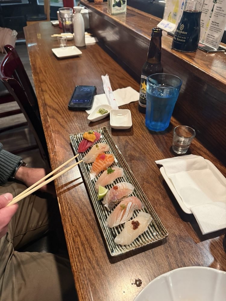 A rectangular plate of assorted sashimi with garnishes, placed on a wooden table in a restaurant. A person is holding chopsticks with sushi pieces. Surrounding items include a smartphone, small dishes with wasabi and soy sauce, a bottle of beer, a gl