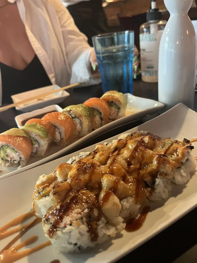 Sushi rolls on a white rectangular plate, topped with eel sauce and mayonnaise, in a restaurant setting with a glass of water, a soy sauce dish, and a person's hand in the background.