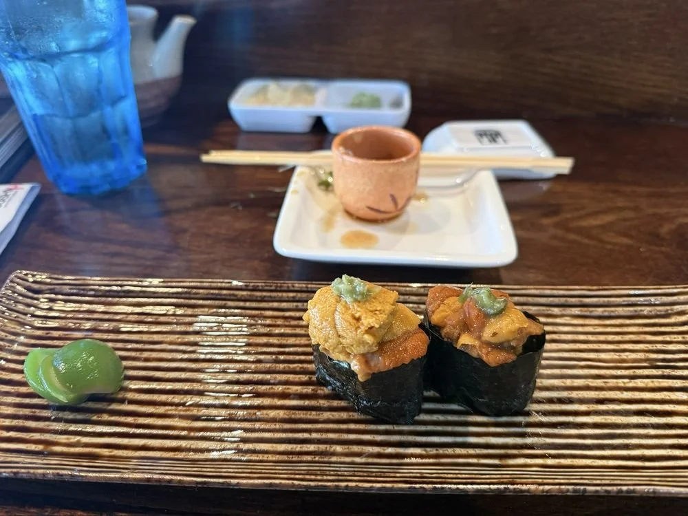 Two pieces of sushi with seaweed, topped with orange fish roe, yellowish sea urchin, and a small amount of green wasabi on top, served on a ceramic plate with a dollop of wasabi on the side.