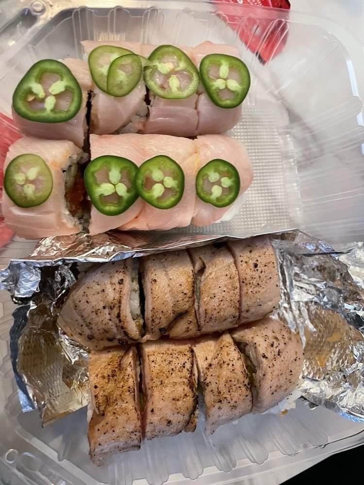 Sushi rolls topped with slices of jalapeño peppers on a clear plastic container with foil-wrapped rolls below, seasoned and cooked chicken wrapped in foil.