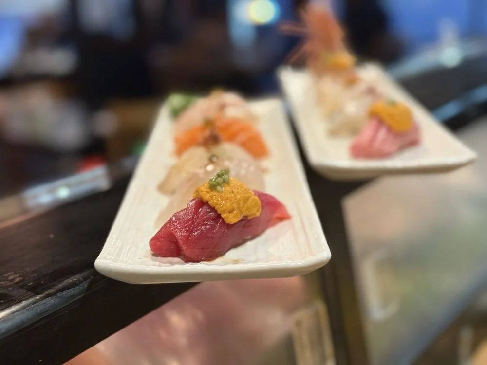 Close-up of two rectangular plates with assorted sushi pieces on a dark counter at a sushi restaurant.